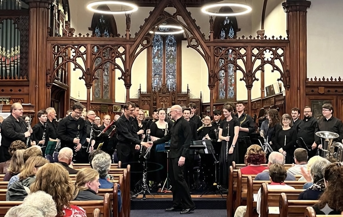 Orchestra in black attire on stage inside a church with wooden arches and stained glass windows, shaking hands with the conductor.