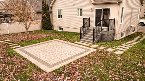 Backyard with a square paved patio, green grass scattered with autumn leaves, a tree with some orange leaves, and a small staircase with black railing leading to a beige house.