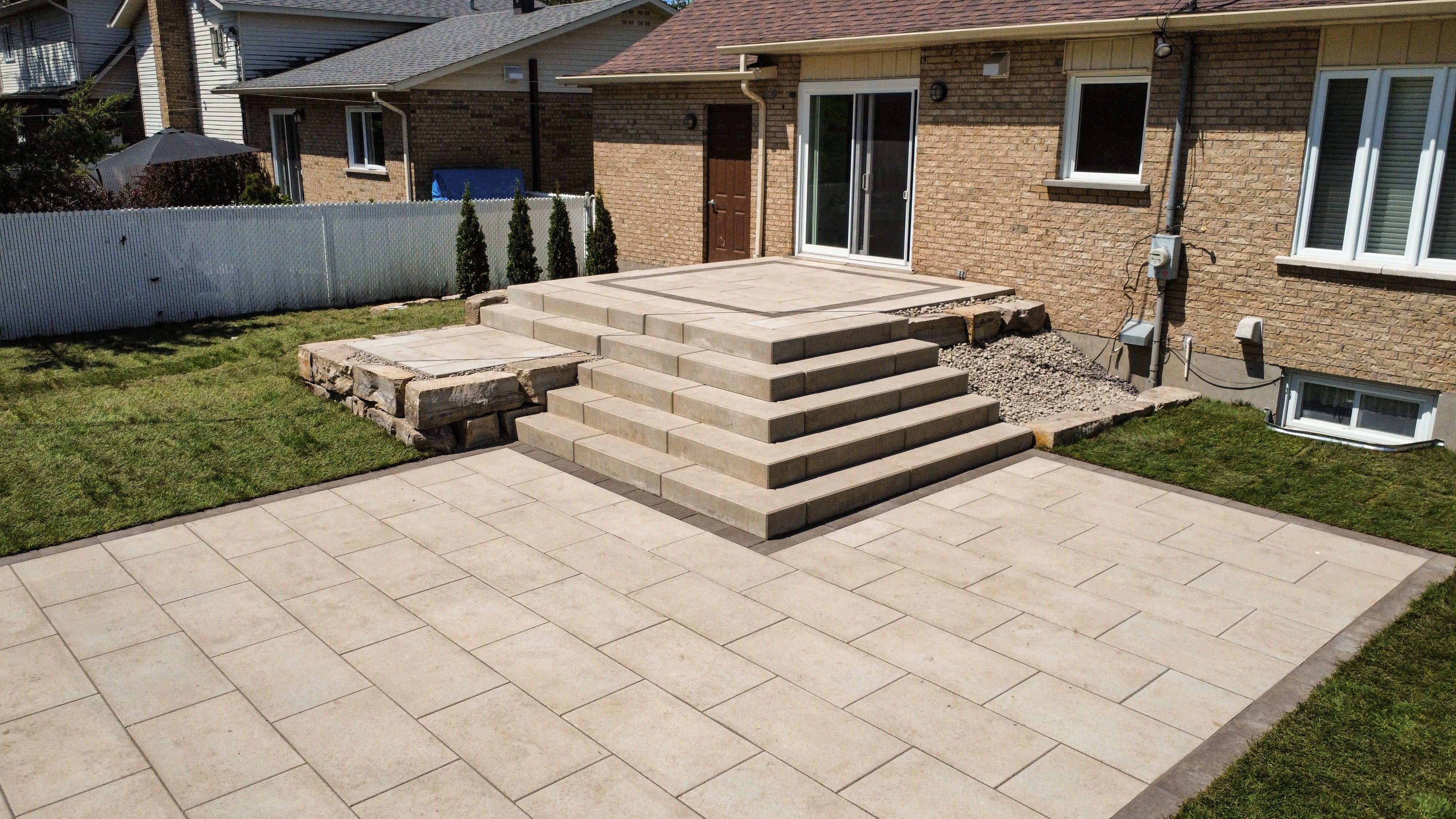 Backyard with a beige tiled patio, wide stone steps leading to a raised platform in front of a brick house with sliding glass doors.