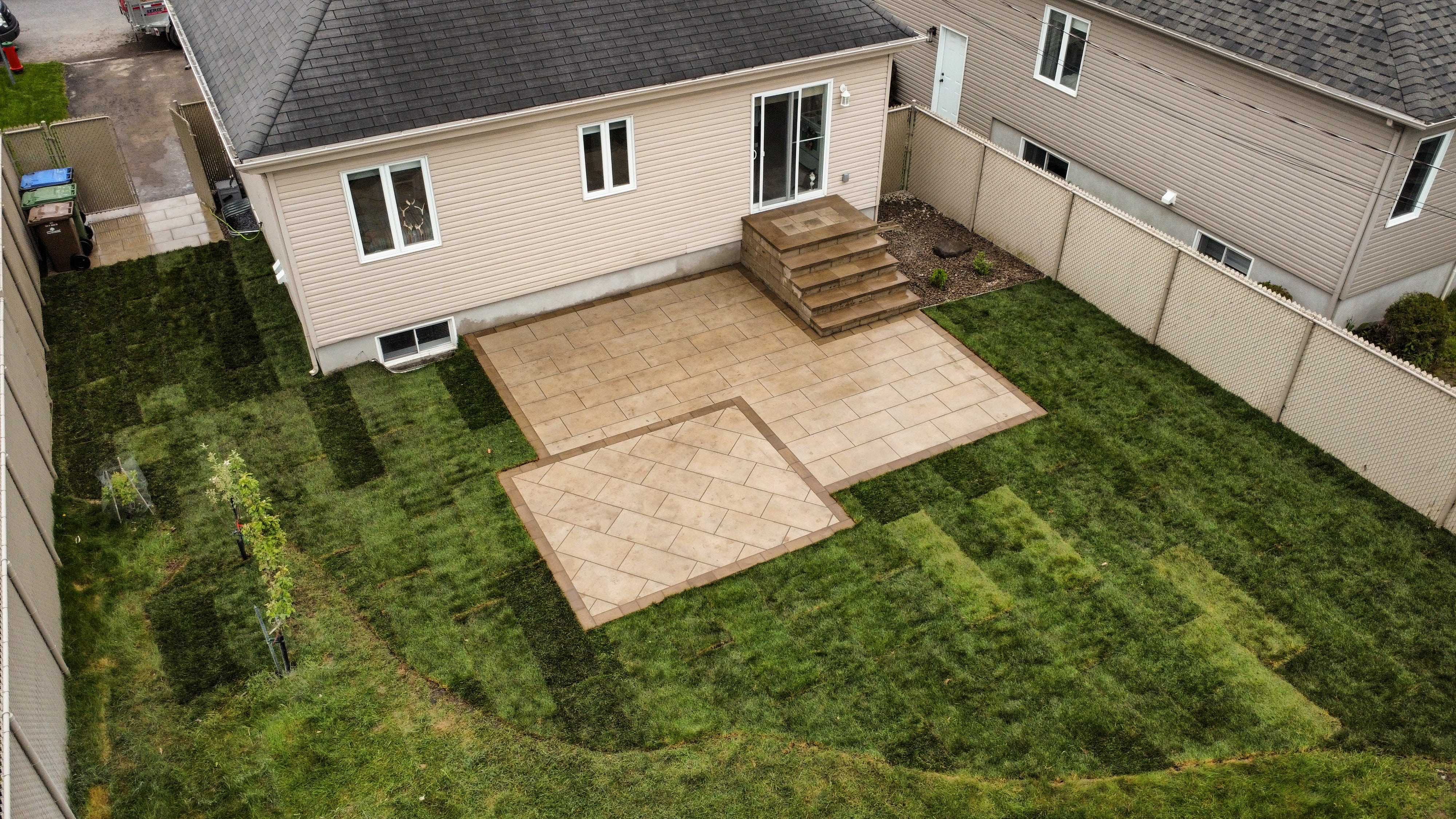 Aerial view of a house backyard with a beige tiled patio, steps leading to a sliding door, green lawn with patchy grass squares, and a fenced perimeter.
