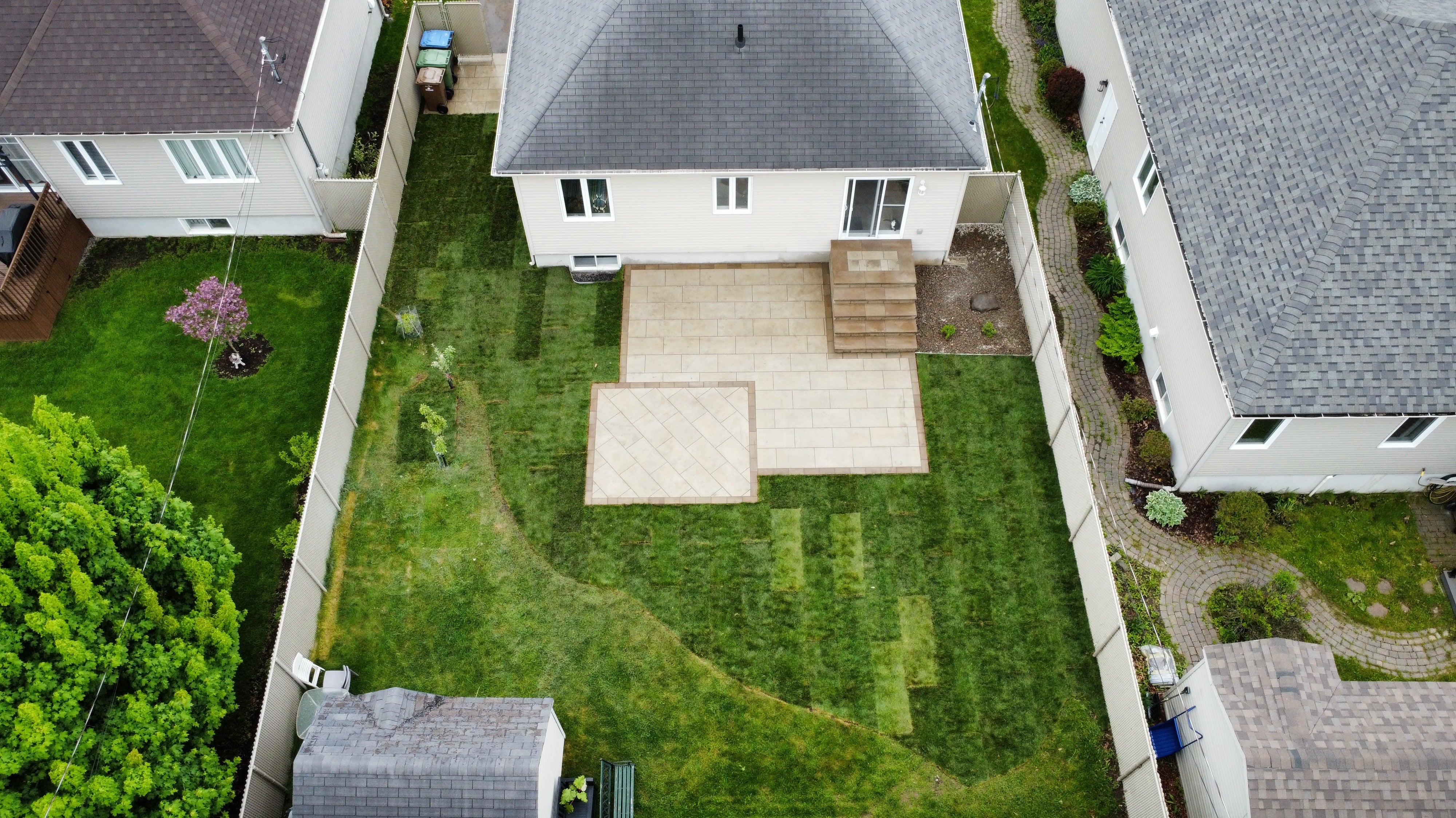 Aerial view of a fenced backyard with a tiled patio, steps leading to a house, and freshly mowed green lawn with trees and garden beds along the edges.