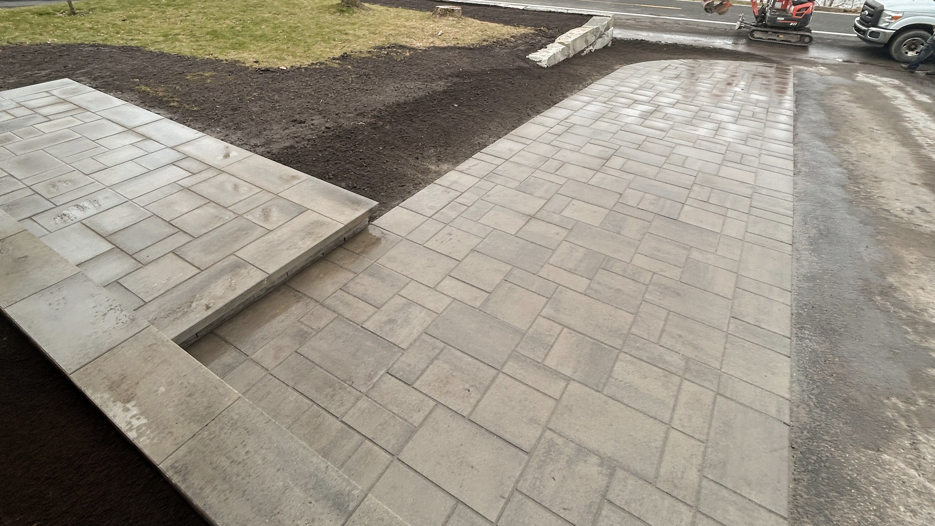 Recently installed beige stone paver driveway and elevated patio with soil and grass surrounding the area.