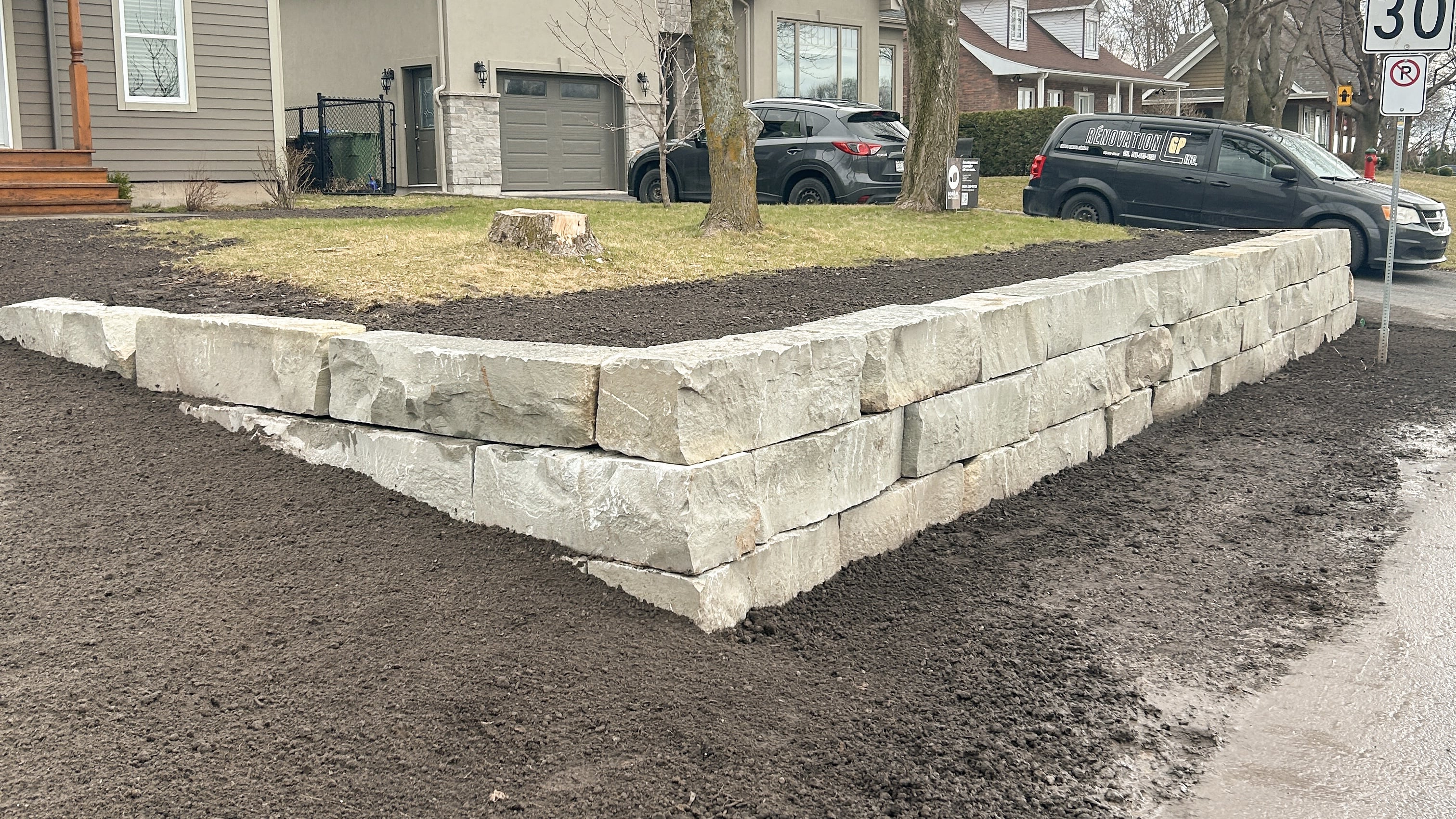 Newly constructed two-tier light gray stone retaining wall around a lawn in front of suburban houses.
