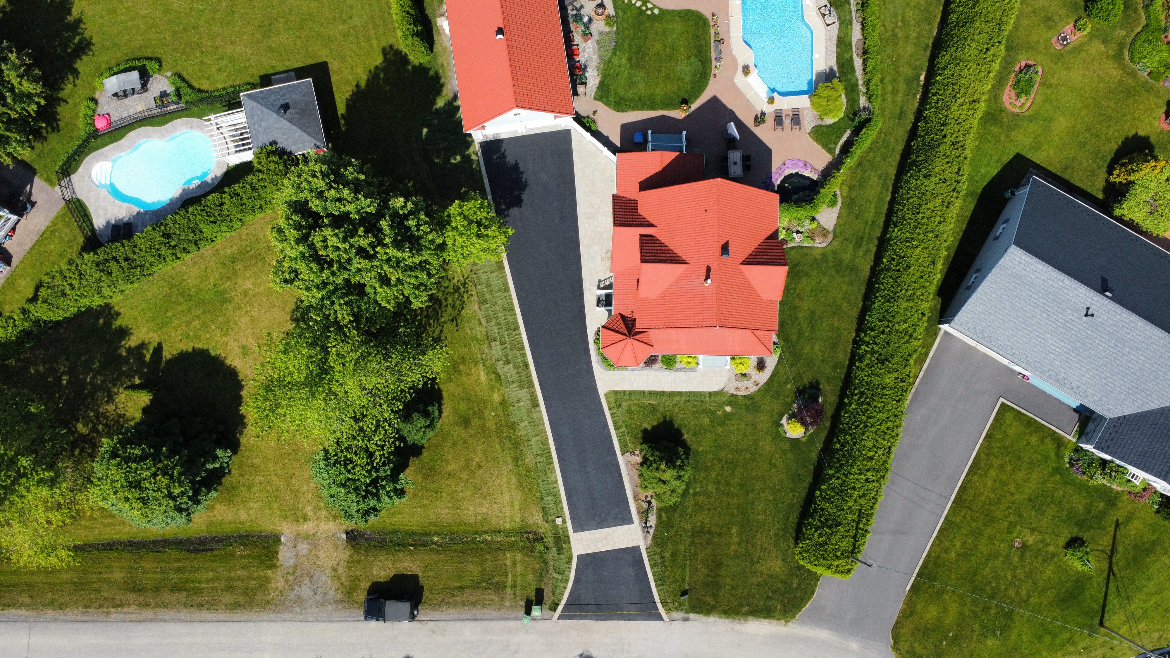 Aerial view of residential houses with red and gray roofs, green lawns, trees, driveways, and two swimming pools.