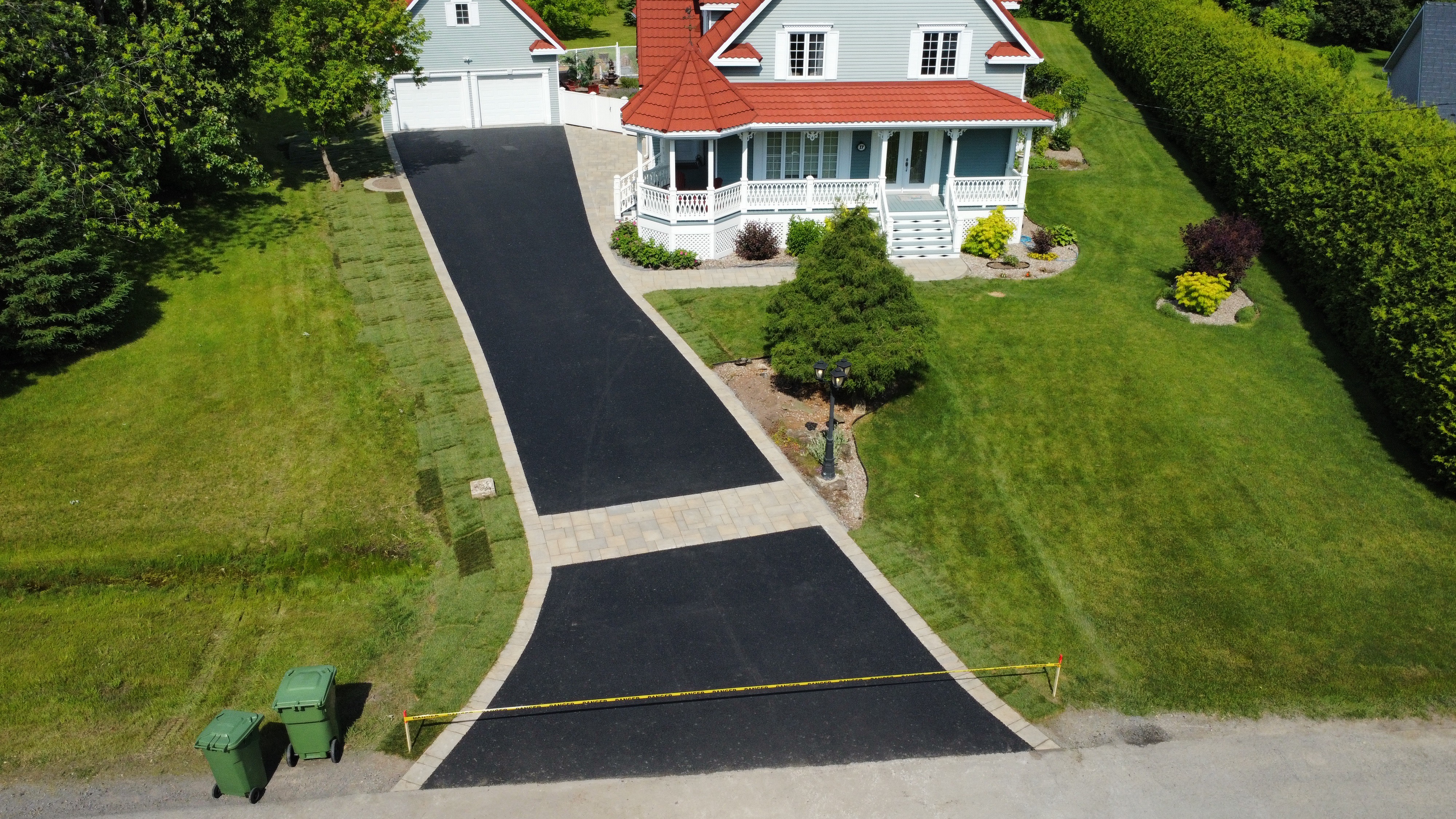 Aerial view of a newly paved black driveway leading to a gray house with red roof, bordered by green lawn and landscaping.