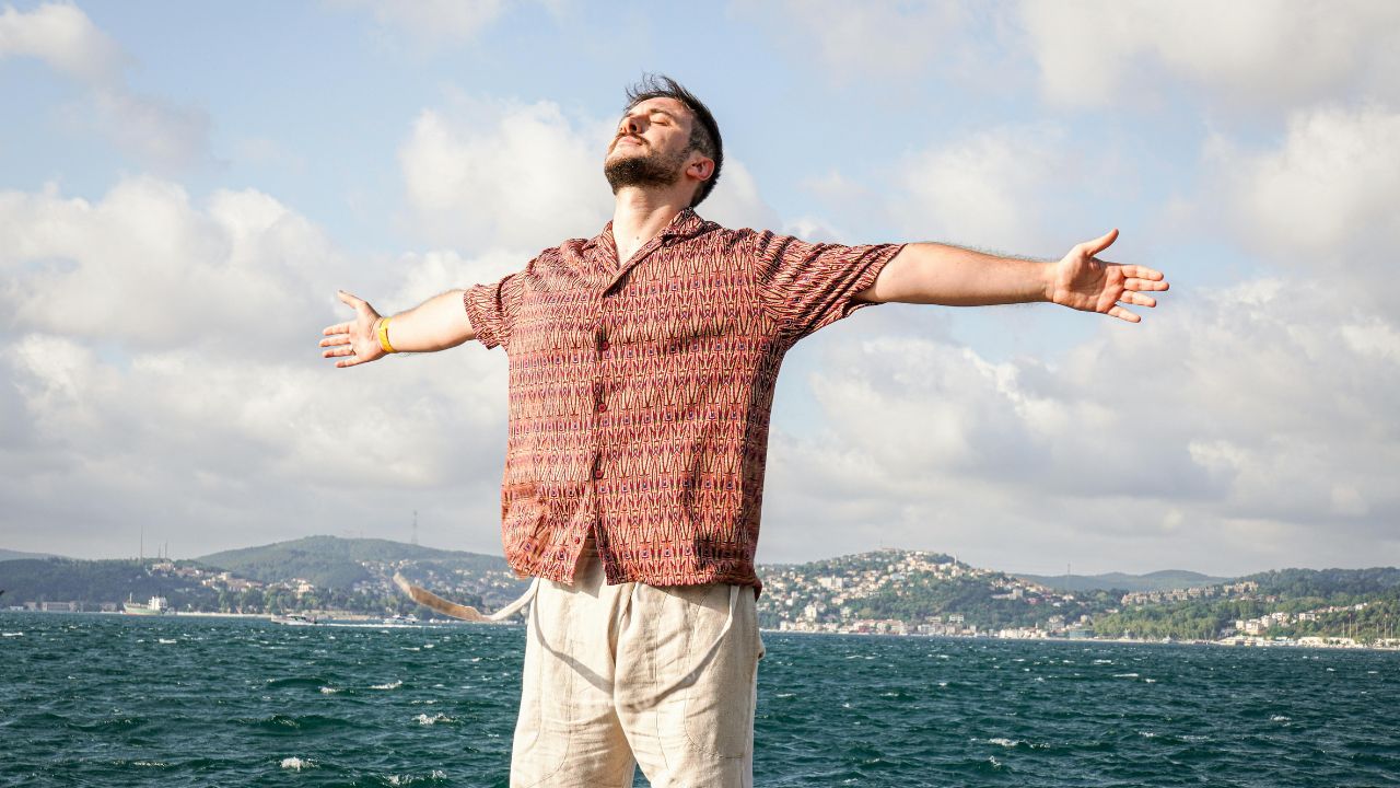 Hombre de pie junto al mar con los brazos abiertos, mirando hacia arriba, disfrutando del viento y el paisaje.