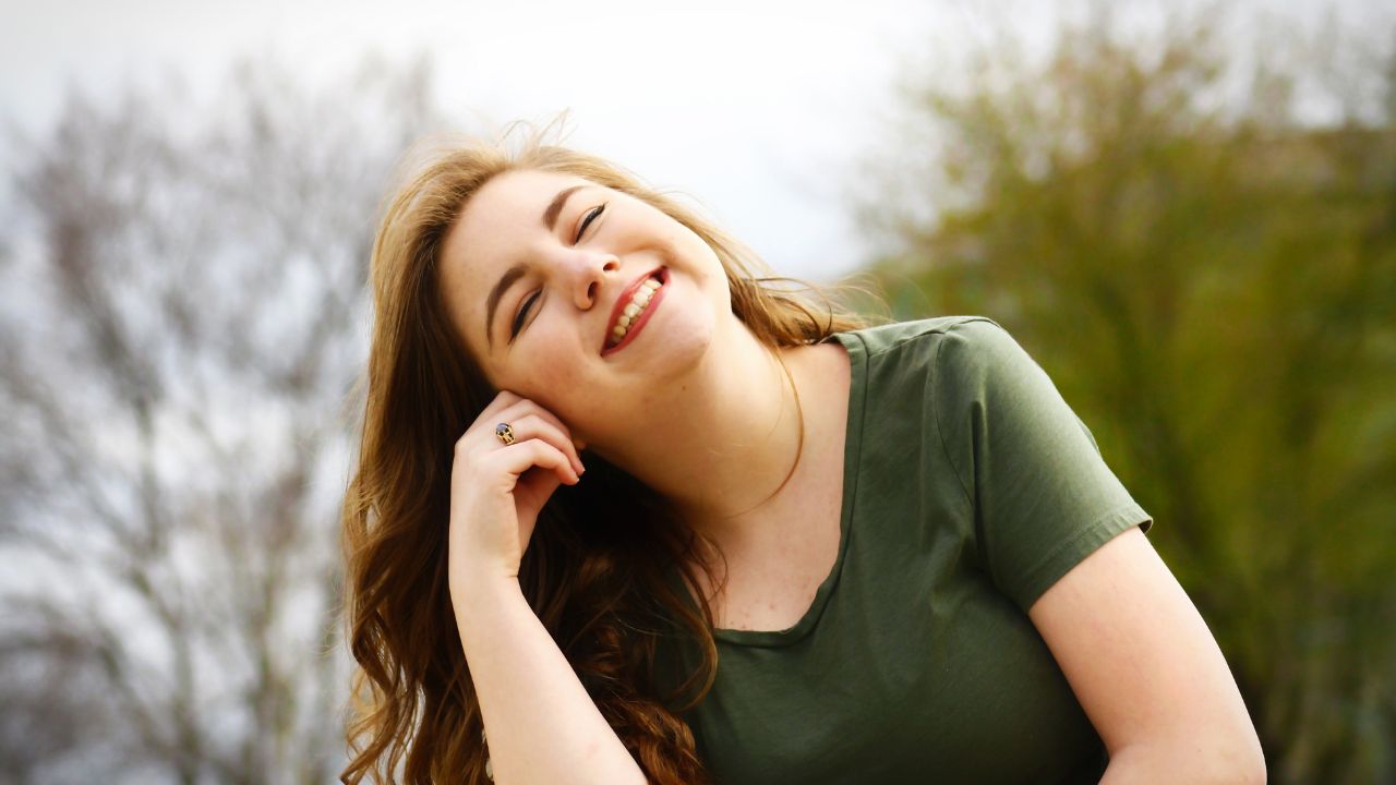 Mujer joven sonriendo al aire libre, con la cabeza ligeramente inclinada.
