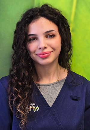 Zu sehen ist eine medizinische Fachangestellte der Praxis. Sie lächelt freundlich und trägt ihre langen dunklen Locken offen. Auf ihrem blauen Arbeitskittel ist das Logo der Gemeinschaftspraxis aufgestickt.