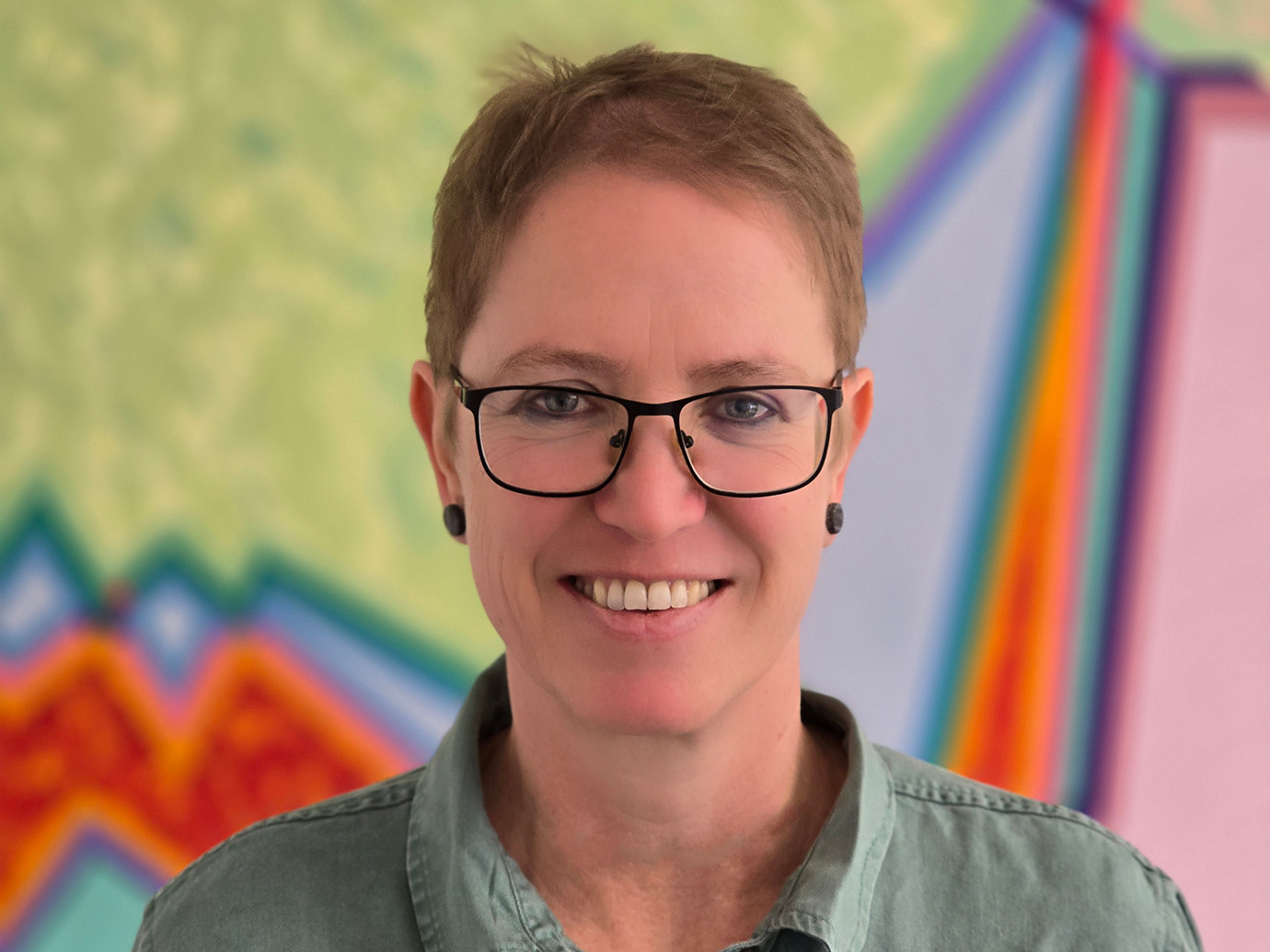 Zu sehen ist Frau Dr. Andrea Müller. Die Brillenträgerin lächelt freundlich. Sie trägt kurzes dunkelblondes Haar. Im Hintergrund ein farbenfrohes abstraktes Gemälde.