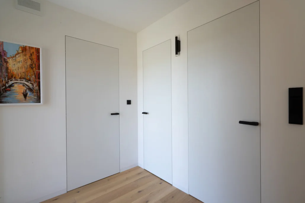 Residential entryway showing three white Fortius doors with concealed door frames and concealed hinges, equipped with black M&T Minimal 2.0 door handles. White oak hardwood flooring.
