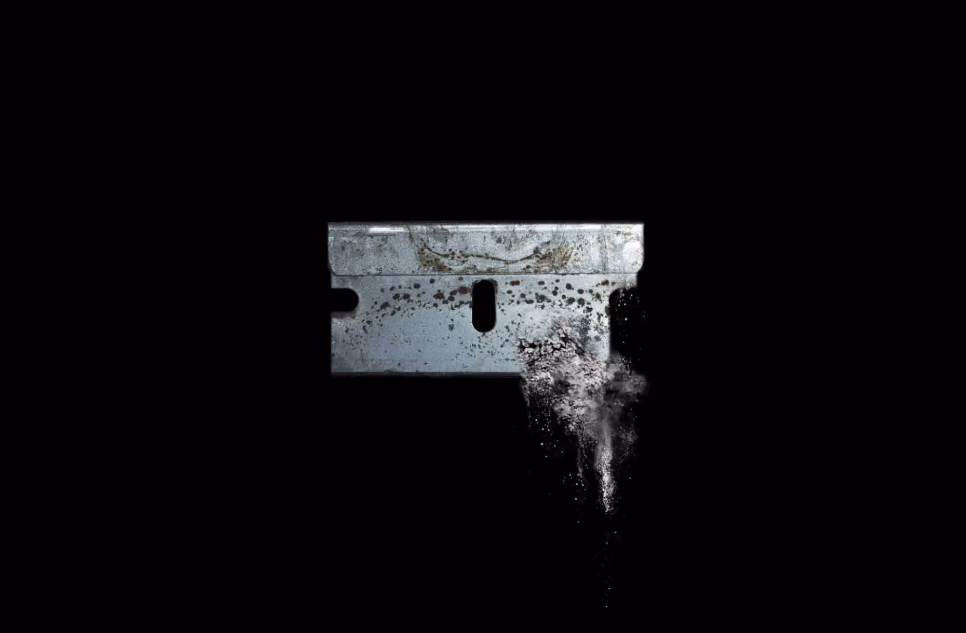 Rusty razor blade with ash or dust particles falling from its edge against a black background.