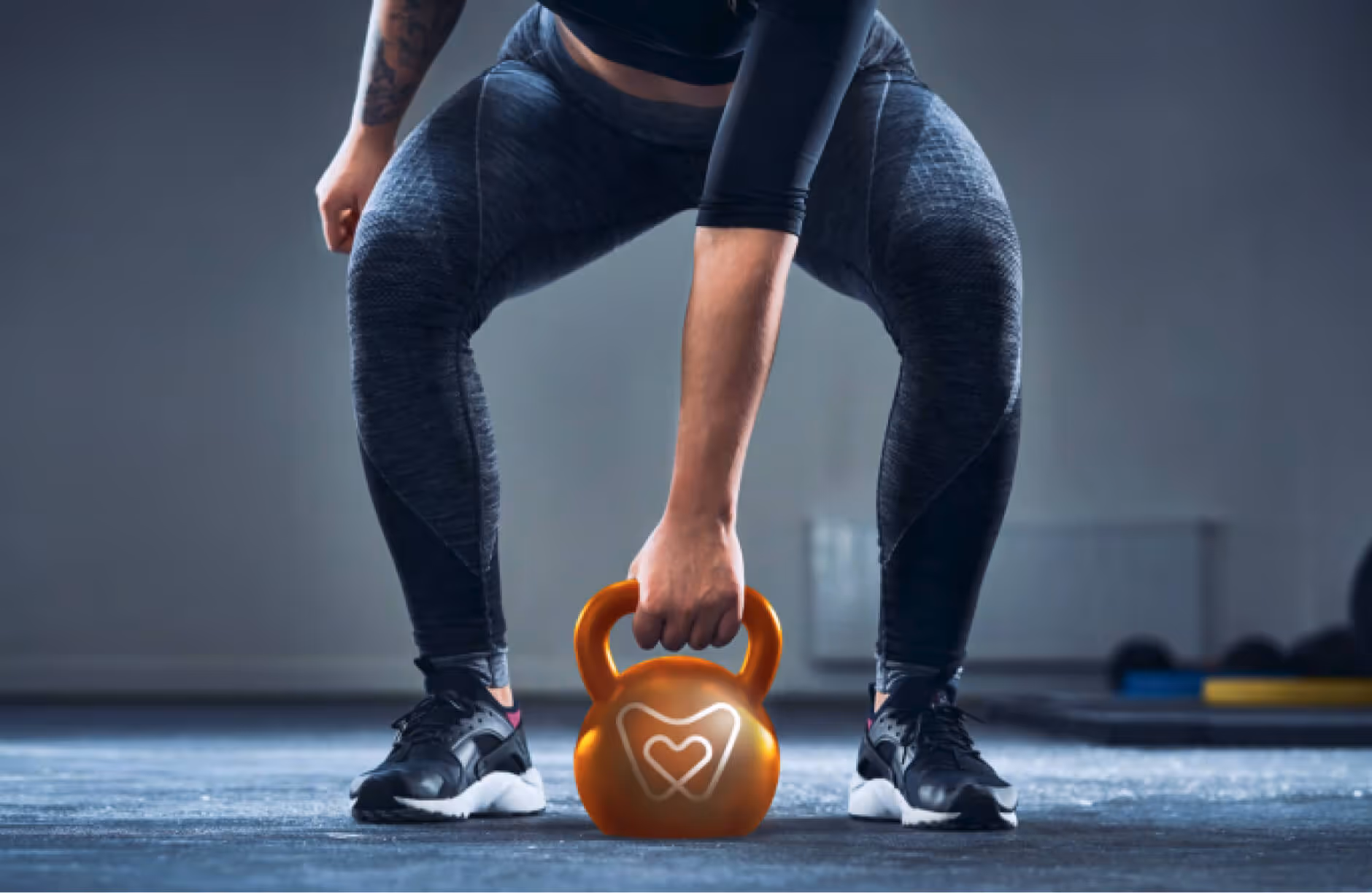 Person in dark athletic wear gripping an orange kettlebell with a heart-shaped design, standing on gym floor.