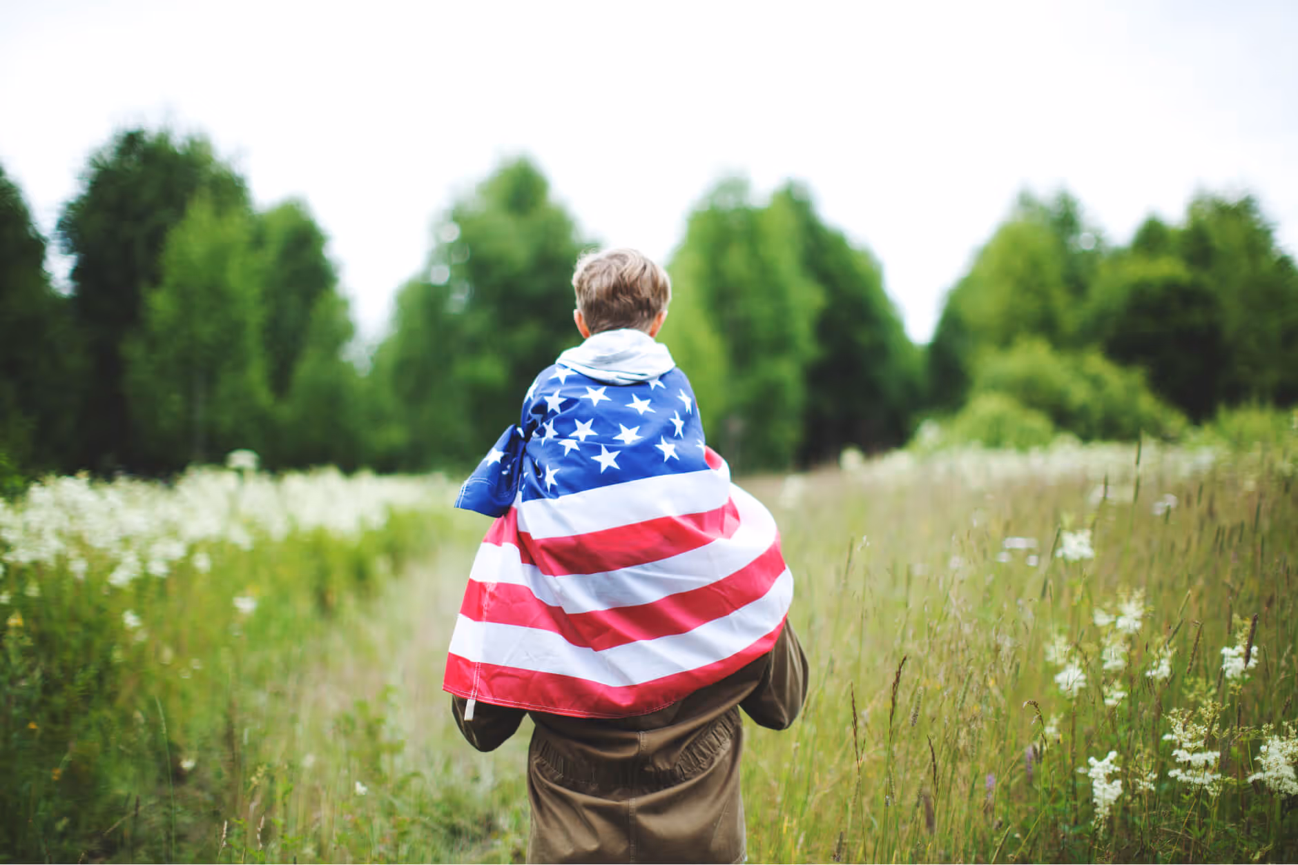 Child wearing an American flag draped over their shoulders, sitting on an adult's shoulders walking along a grassy path with trees in the background.