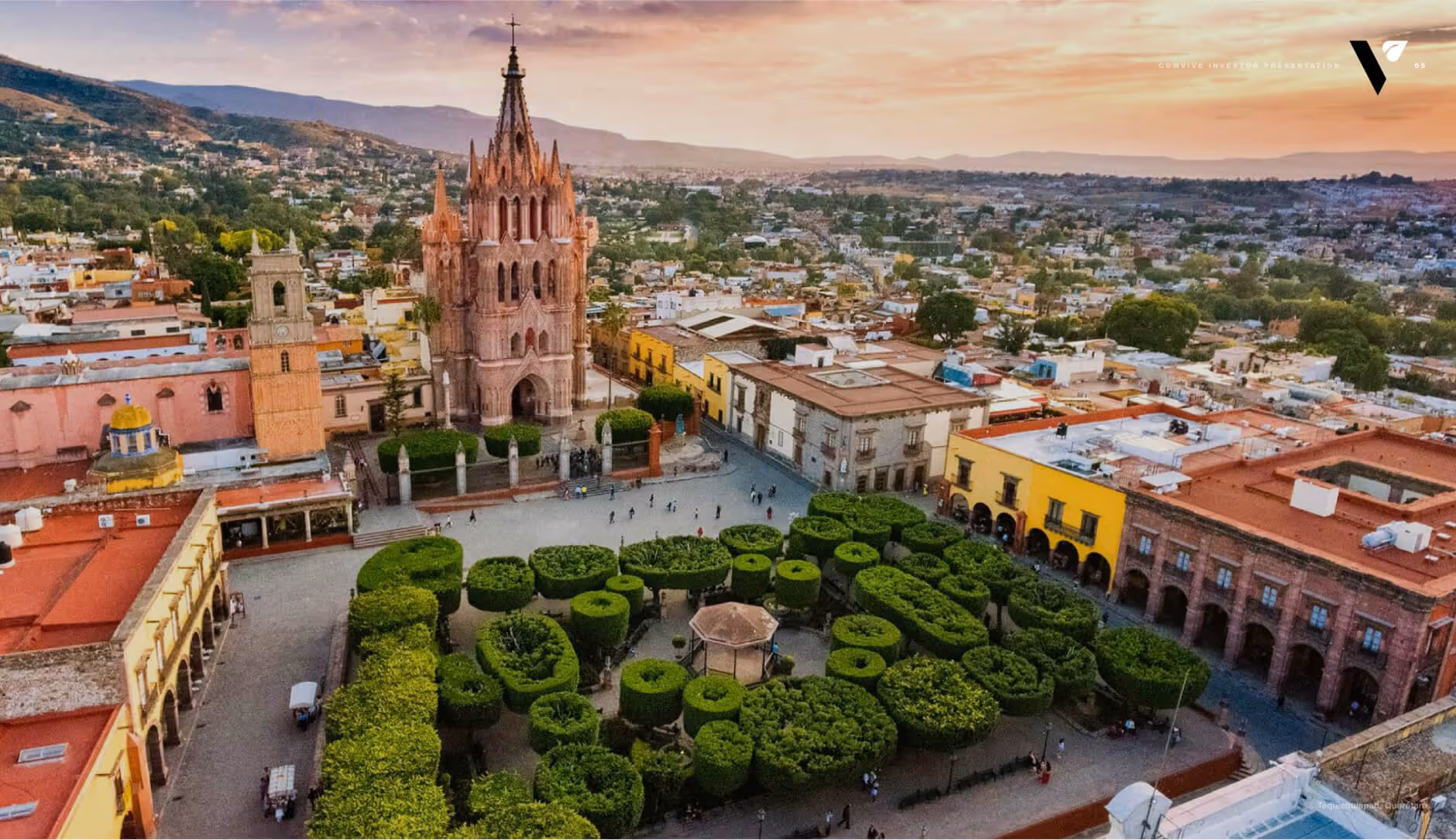 Aerial view of a historic city square with symmetrical green garden, ornate church with tall spires, and surrounding colorful buildings at sunset.