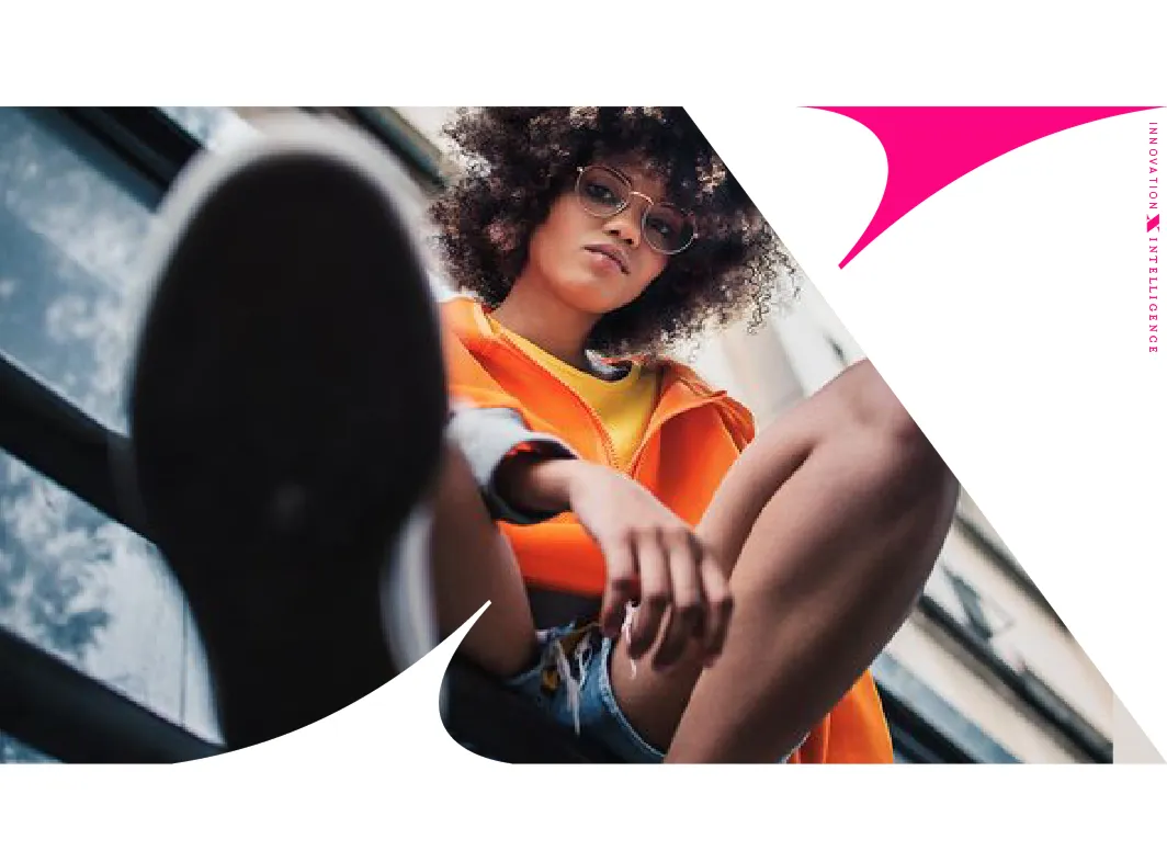 Low-angle photo of a young woman with curly hair and glasses wearing an orange jacket and denim shorts, with one foot close to the camera lens.