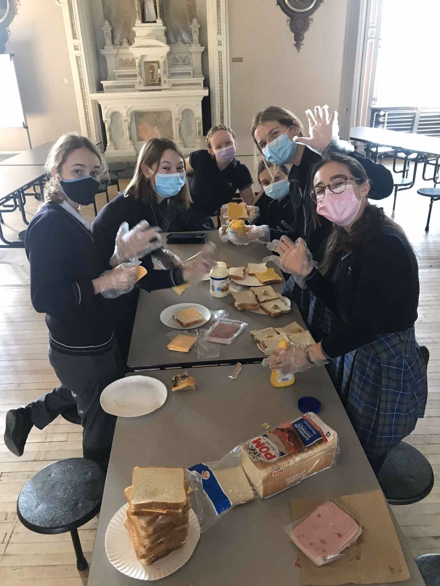 Six masked students wearing gloves prepare sandwiches with bread, cheese, and lunch meat at a long table in a large room.