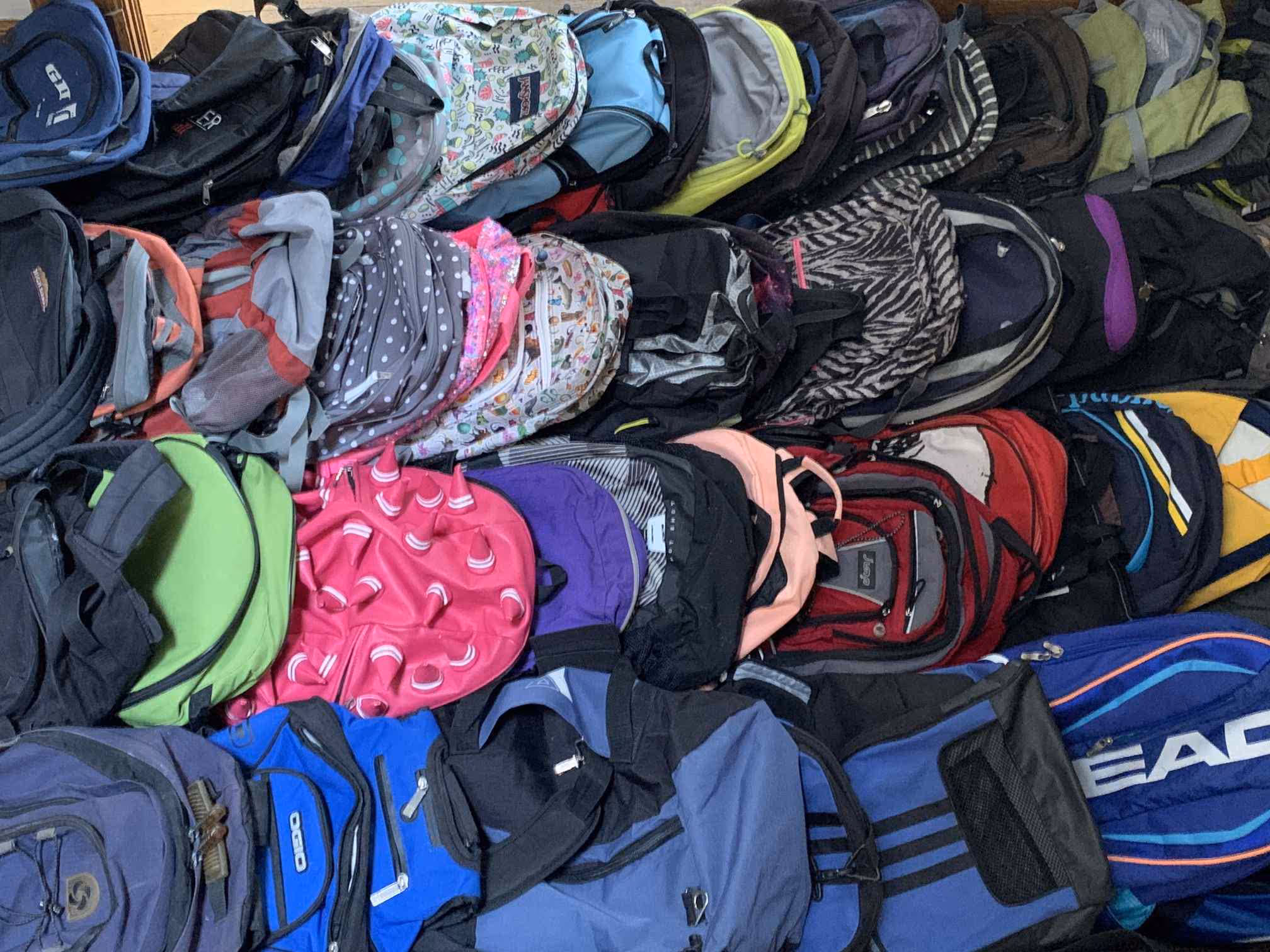 A large pile of assorted backpacks in various colors, patterns, and sizes stacked closely together.