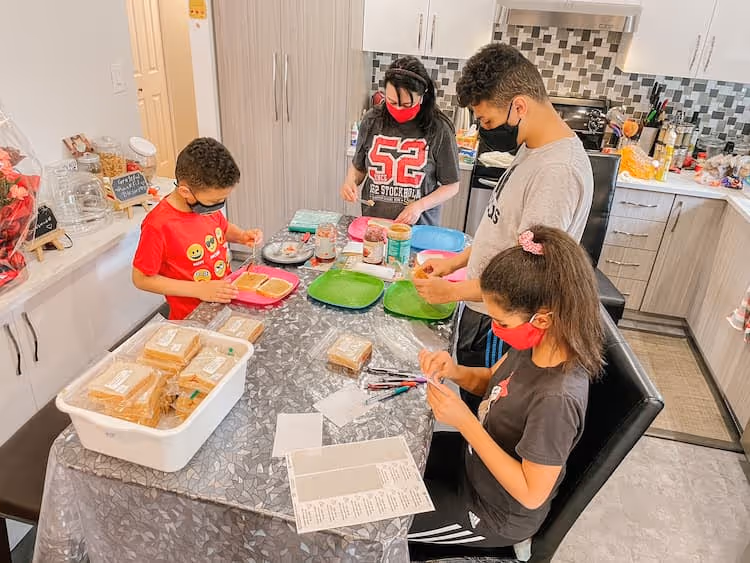 Four children wearing face masks prepare sandwiches together at a kitchen table with plates, bread, and condiments.
