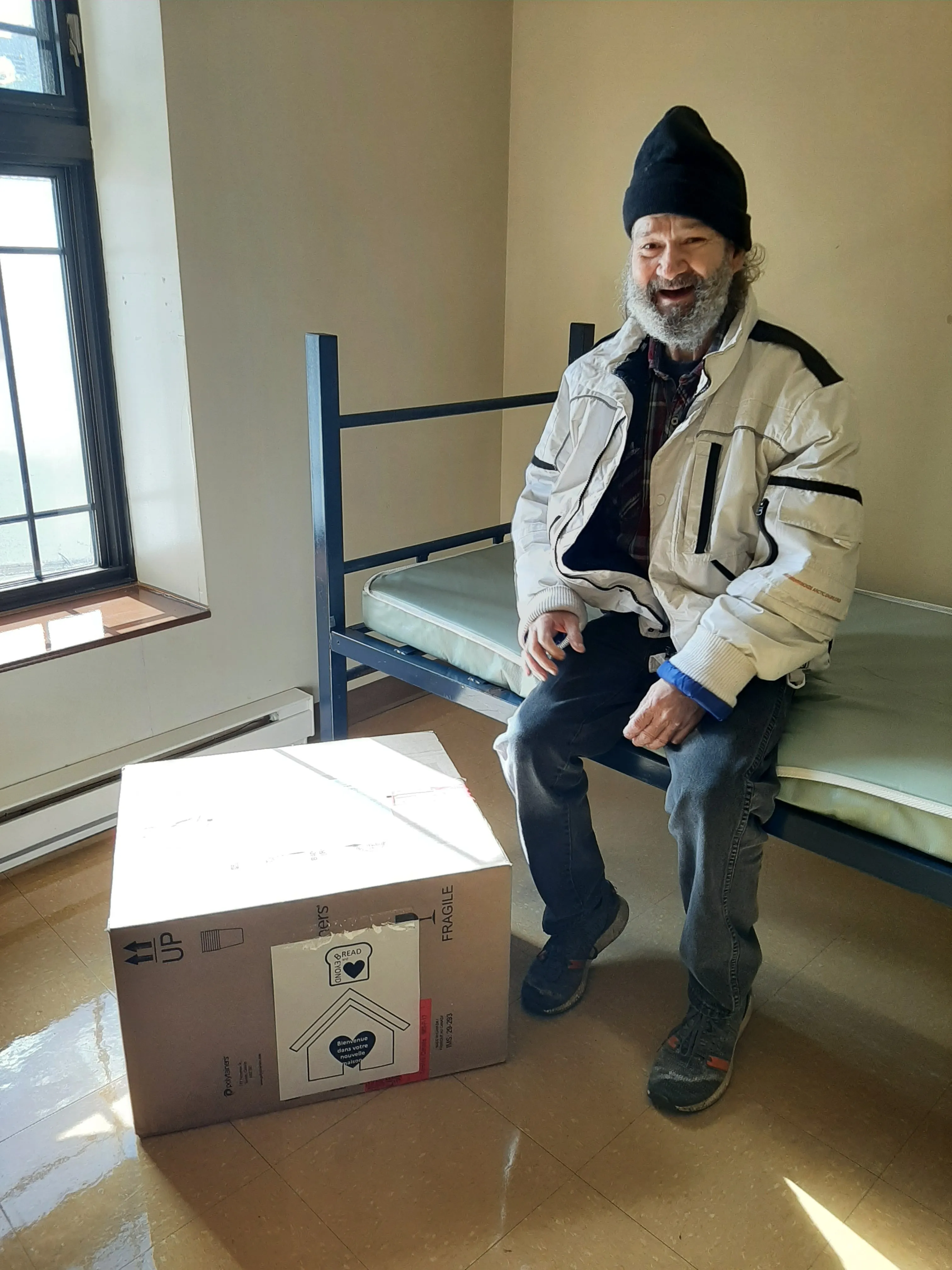 Smiling man with beard and beanie sitting on a metal-framed bed in a sunlit room beside a large cardboard box labeled fragile.