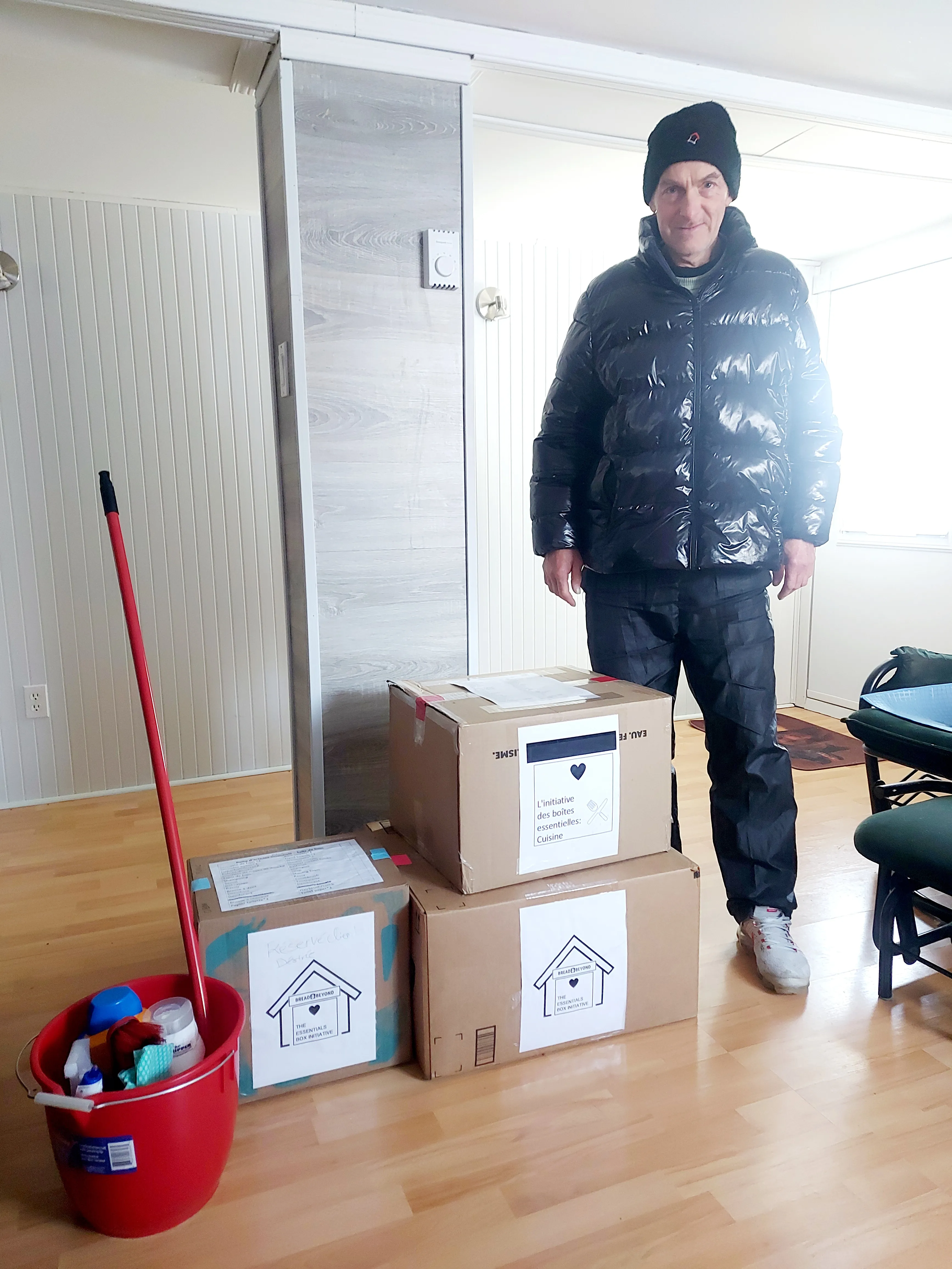 Man wearing black puffer jacket, black hat, and pants standing indoors next to three stacked cardboard boxes labeled 'The Essentials Box Initiative' and a red bucket with cleaning supplies and a mop.