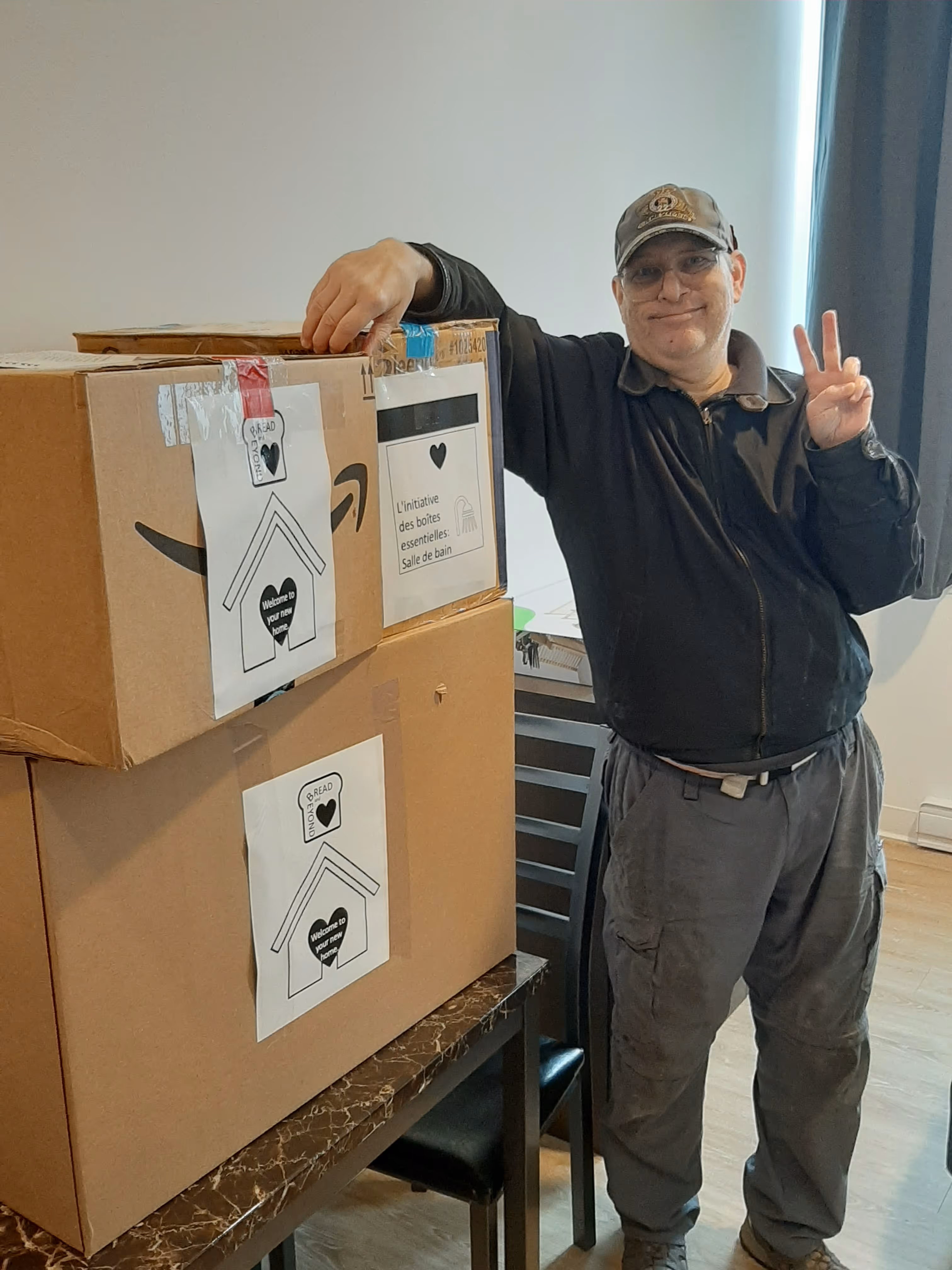 Smiling man wearing a cap and glasses stands next to stacked cardboard boxes with welcome labels, making a peace sign.