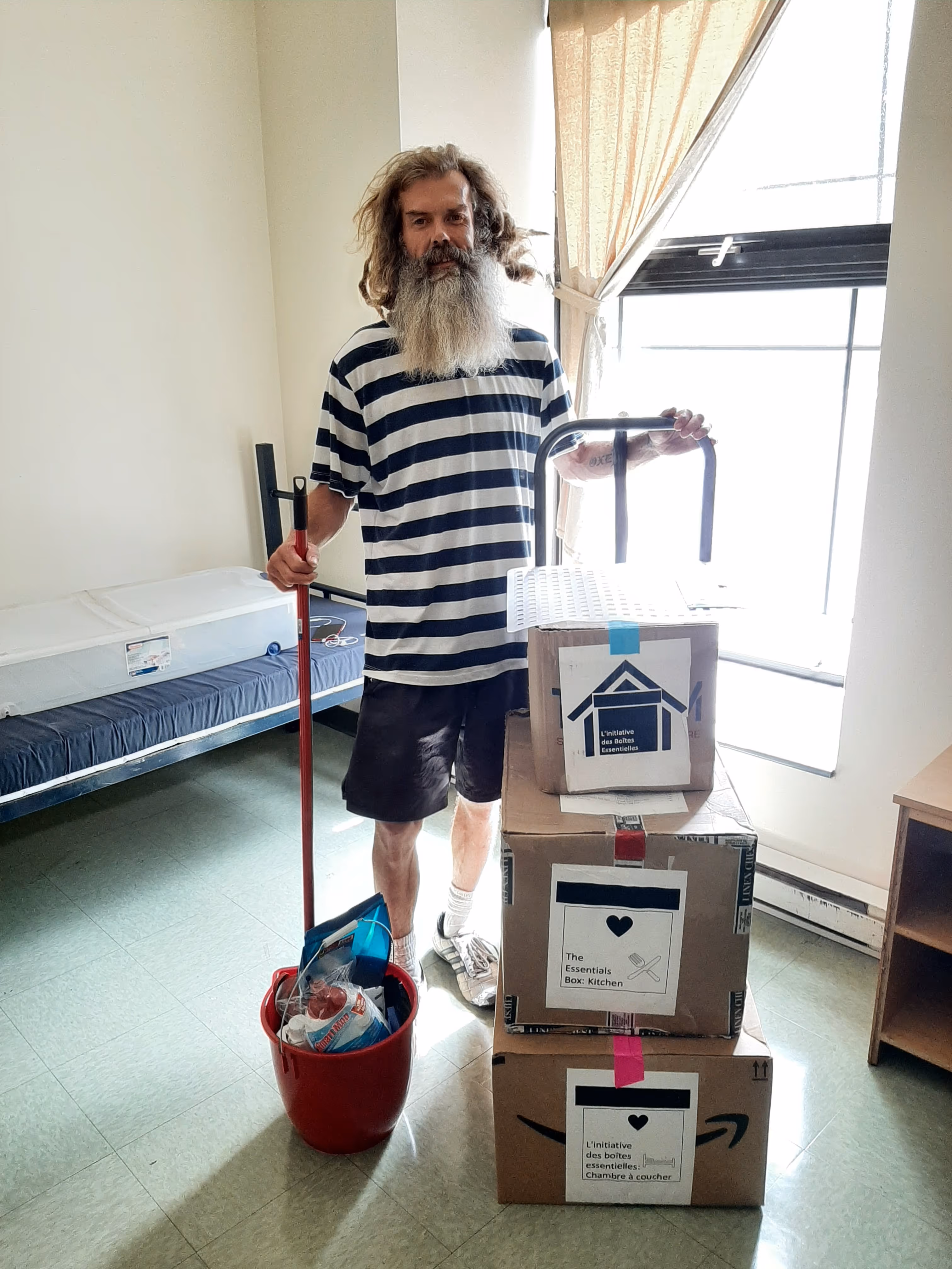 Man with long hair and beard holding a mop in a red bucket next to a stack of three labeled cardboard boxes in a sparsely furnished room.