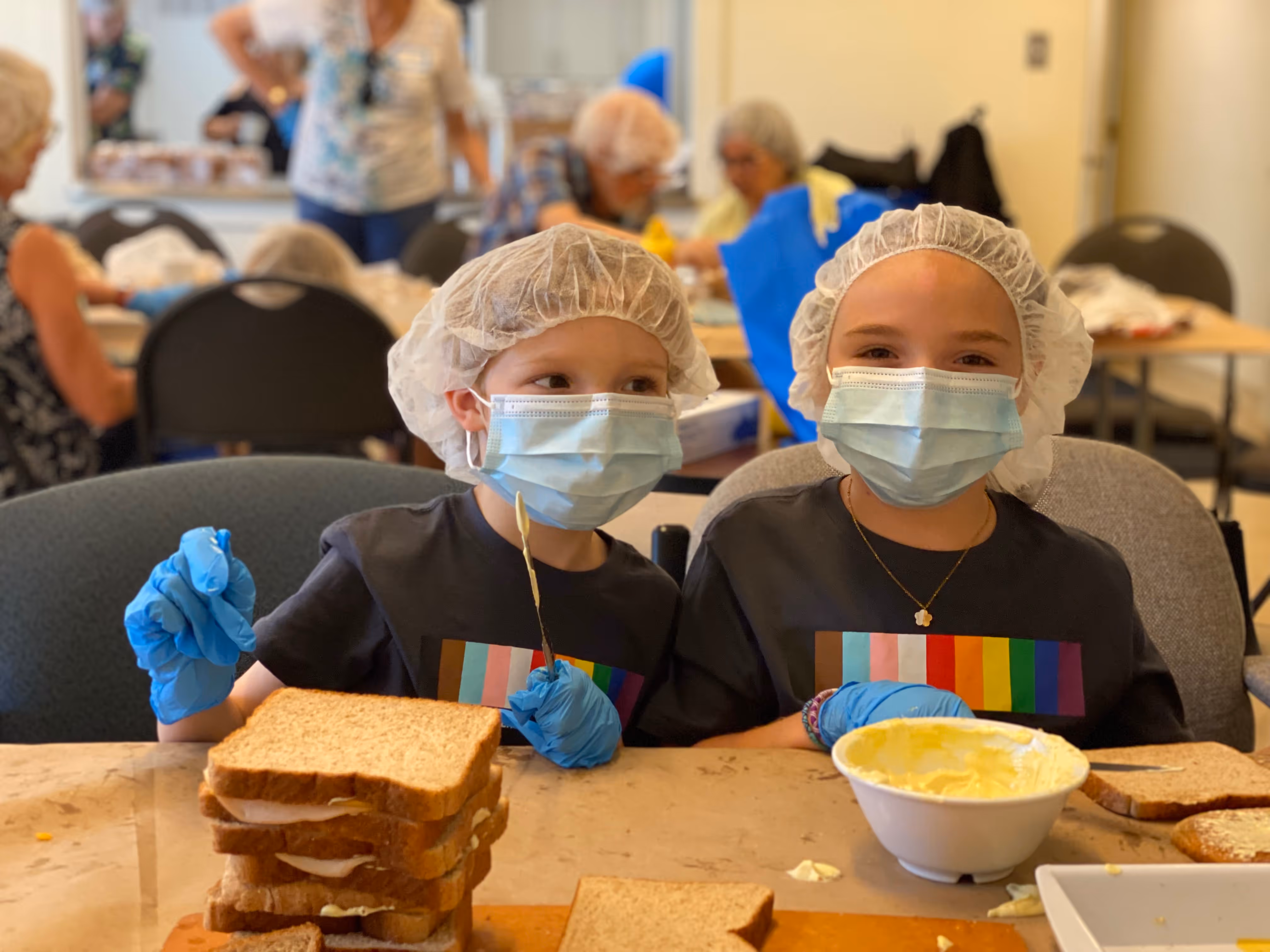 Two children wearing hairnets, face masks, and gloves making sandwiches at a table.