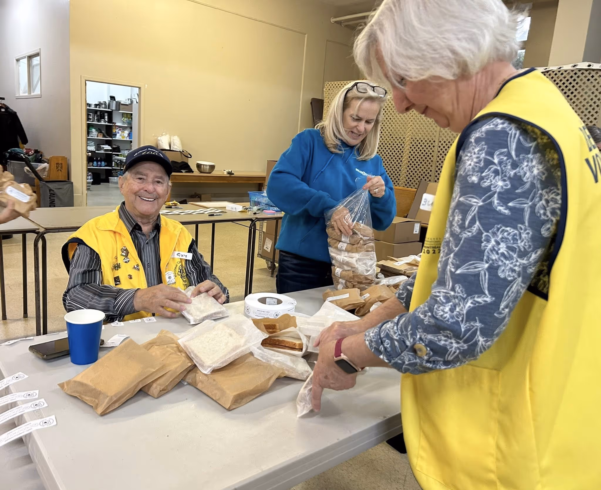 Three volunteers packaging sandwiches at a table inside a community center.