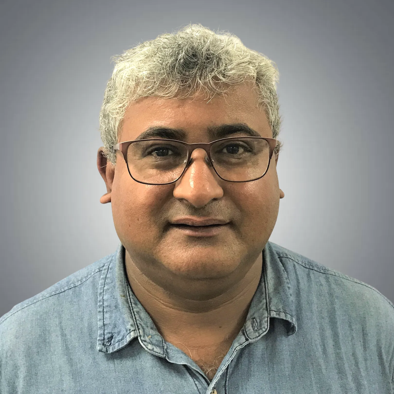 Middle-aged man with salt-and-pepper hair and glasses, wearing a light blue denim shirt, against a gray gradient background.