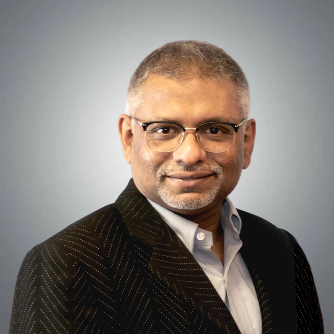 Portrait of a middle-aged man with gray hair and glasses wearing a black blazer and light blue shirt against a gray background.