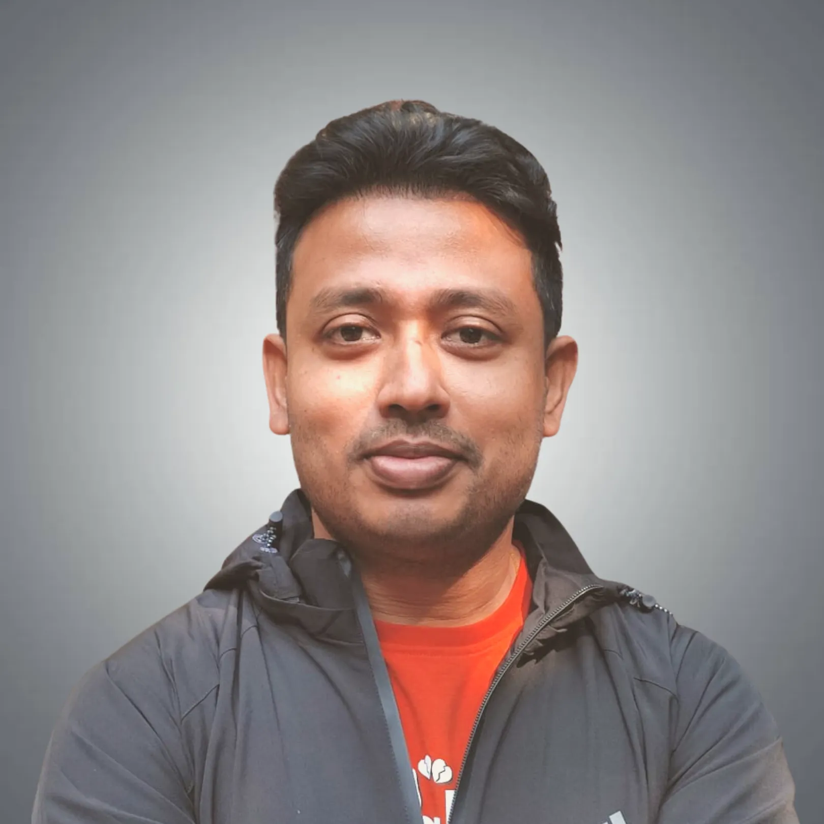 Portrait of a man with short black hair wearing a black jacket and an orange shirt against a gray gradient background.