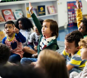 Diverse group of young children sitting on the floor in a classroom, with two children raising their hands.