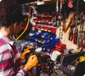 Mechanic wearing orange gloves working on a vehicle part in a cluttered workshop with tools and containers on shelves.