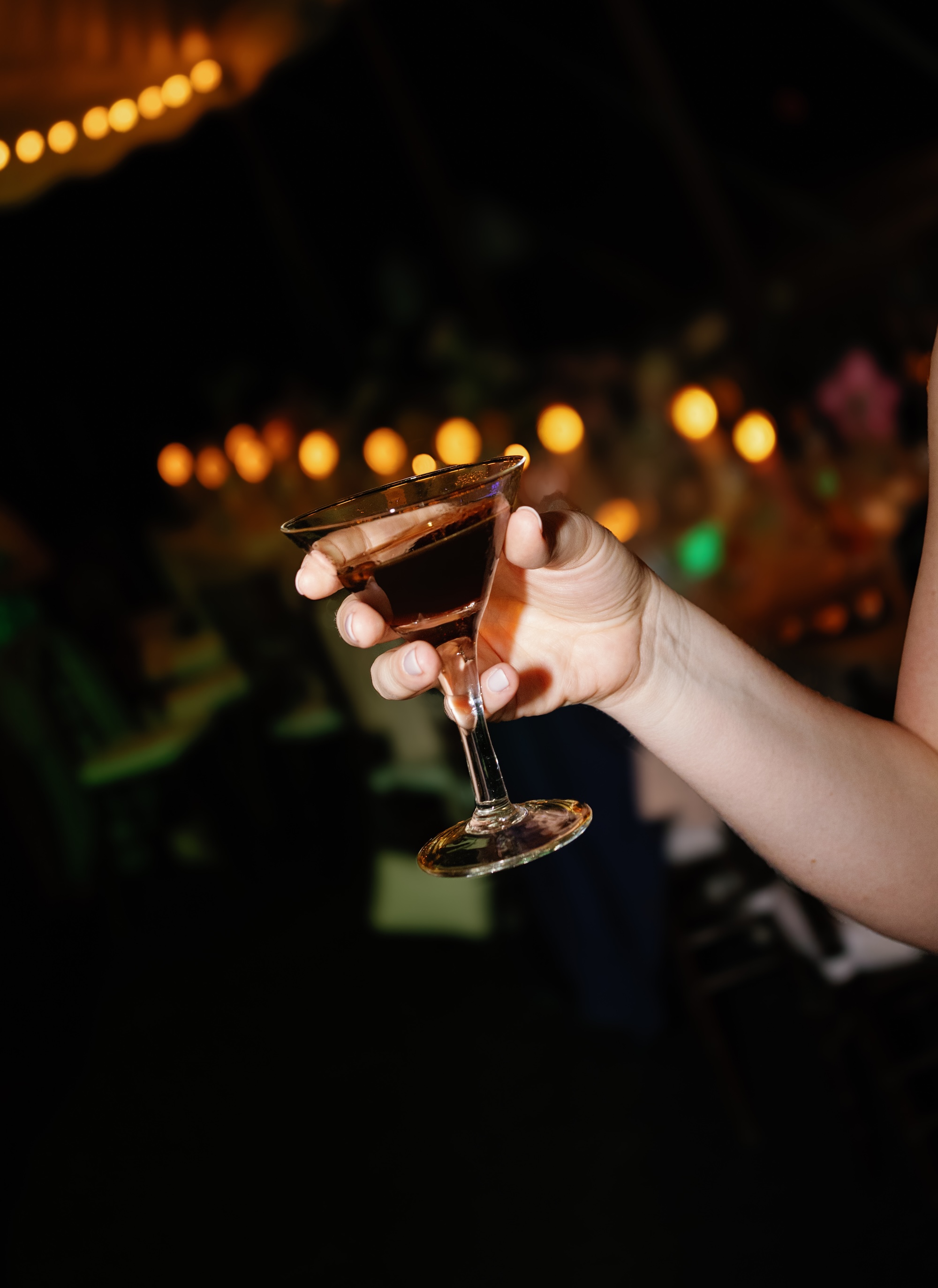 A hand holding a tilted espresso martini in a dimly lit ballroom with sparkling string lights in the background.