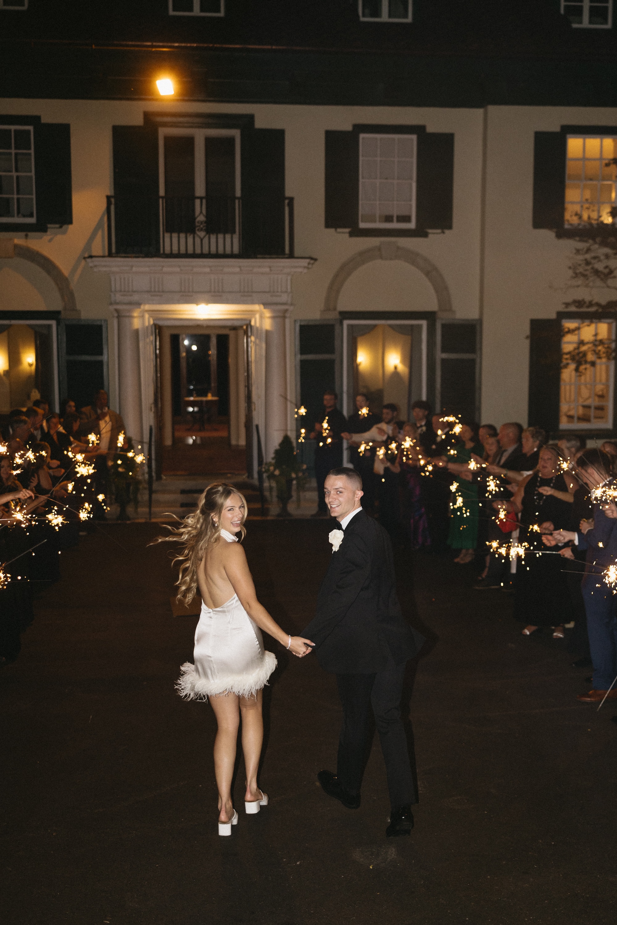 A bride in a short feathered dress and groom in a suit walking toward the mansion entrance, with guests alongside them and sparklers lighting the path.