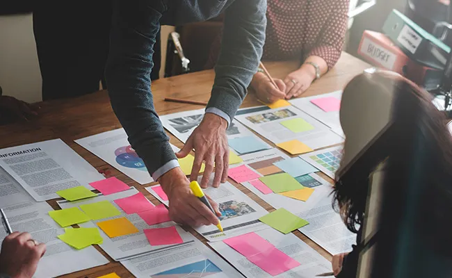 Un groupe de personnes collabore autour d’une table recouverte de documents et de post-its colorés. Une personne se penche et utilise un surligneur jaune sur un papier. La scène suggère une séance de brainstorming ou de planification dans un cadre professionnel.