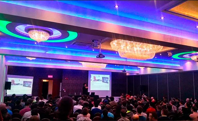 Salle de conférence moderne éclairée en bleu avec un orateur présentant devant un public nombreux.