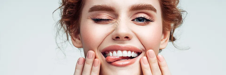 Gros plan d'une personne souriante aux cheveux roux ondulés, à la peau bronzée et aux yeux bleus. Ils clignent d’un œil et tirent de manière ludique leur langue entre leurs dents tout en tenant leur visage à deux mains, montrant des ongles soignés et manucurés.