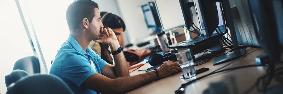Un homme en chemise bleue est assis à un bureau, regardant un écran d'ordinateur avec sa main posée sur son menton. Une autre personne, partiellement floue, travaille également sur un ordinateur en arrière-plan. Le décor semble être un bureau ou un espace de travail.