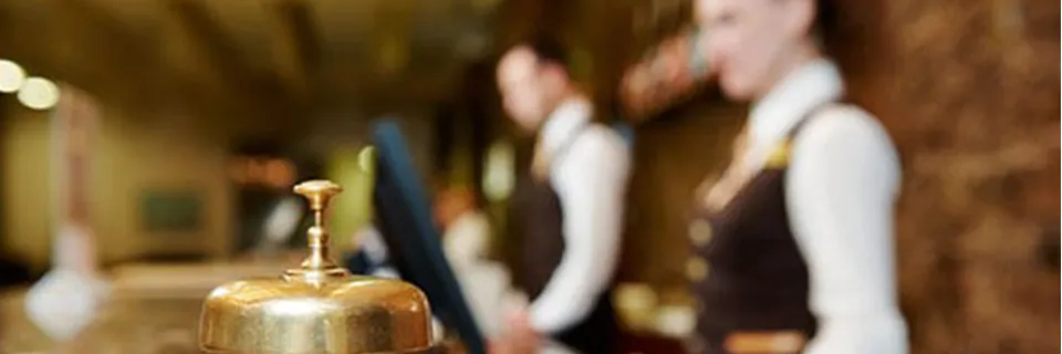 Un gros plan d’une cloche de service dorée sur la réception d’un hôtel. À l’arrière-plan, un homme et une femme du personnel de l’hôtel en uniforme se tiennent au bureau, légèrement flous. Le cadre bénéficie d'un éclairage chaleureux et d'une ambiance accueillante.