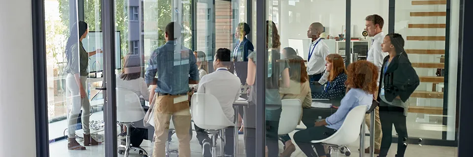 Un groupe de personnes est rassemblé dans une salle de conférence moderne aux murs de verre, écoutant attentivement quelqu'un qui présente à l'avant. Ils sont assis autour d'une table, quelques-uns debout observant la présentation affichée sur un écran.