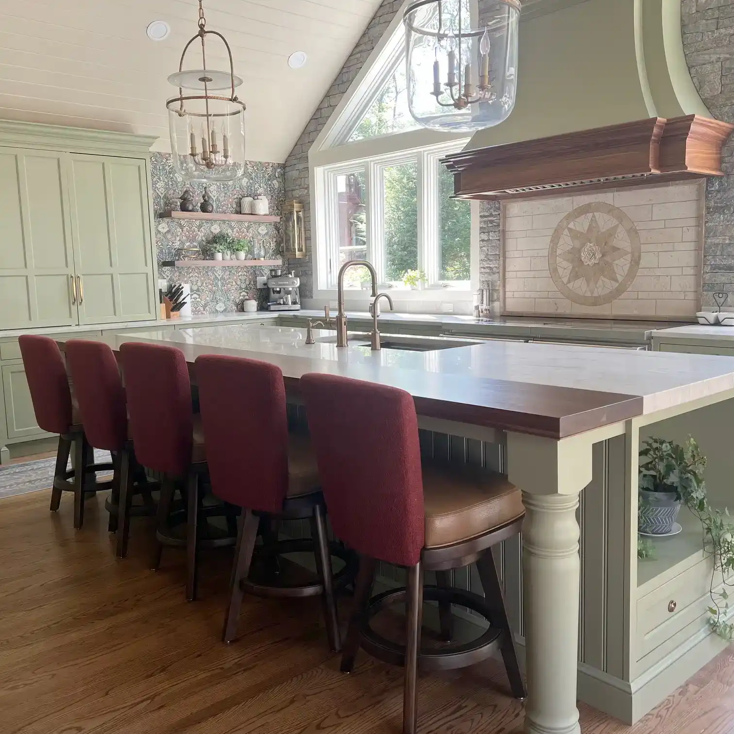 A stylish kitchen features a large island with five red-backed bar stools, light green cabinetry, wood floors, open shelves, and a large window.