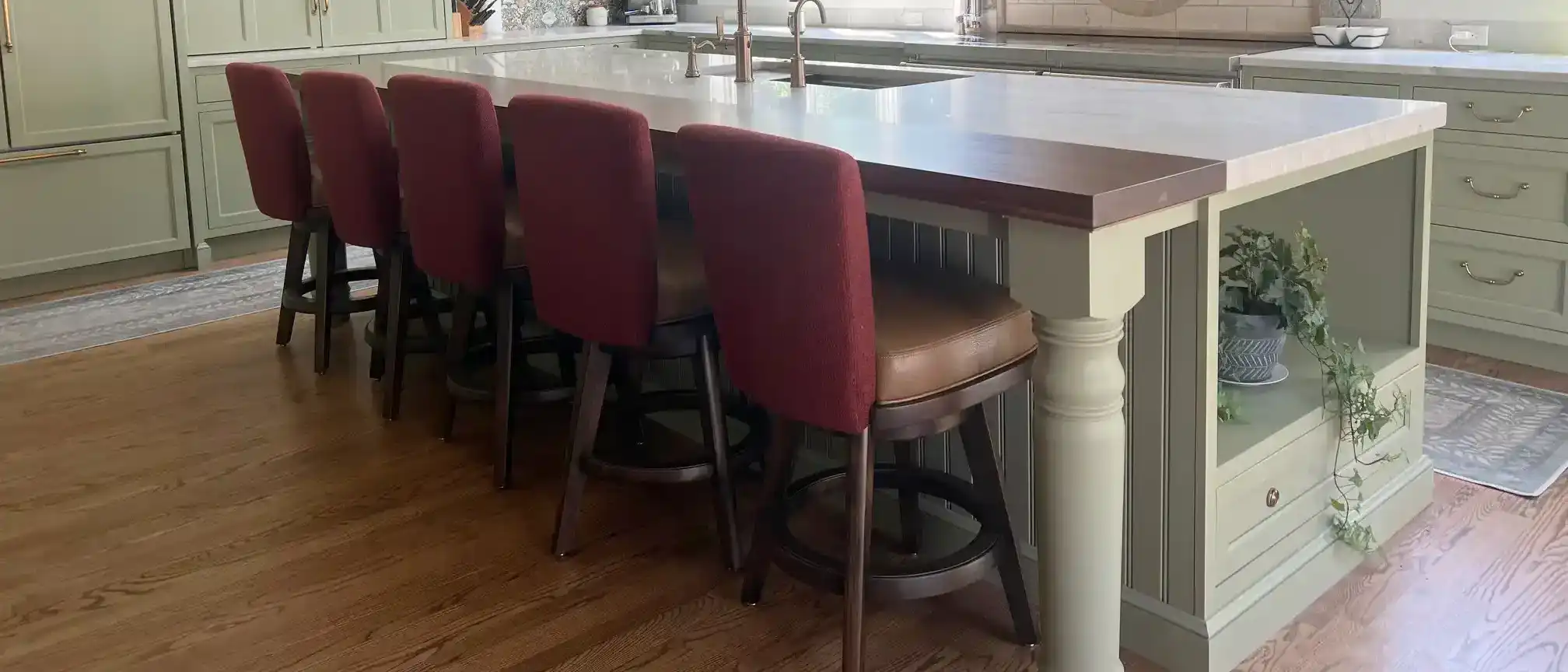 A stylish kitchen features a large island with five red-backed bar stools, light green cabinetry, wood floors, open shelves, and a large window.