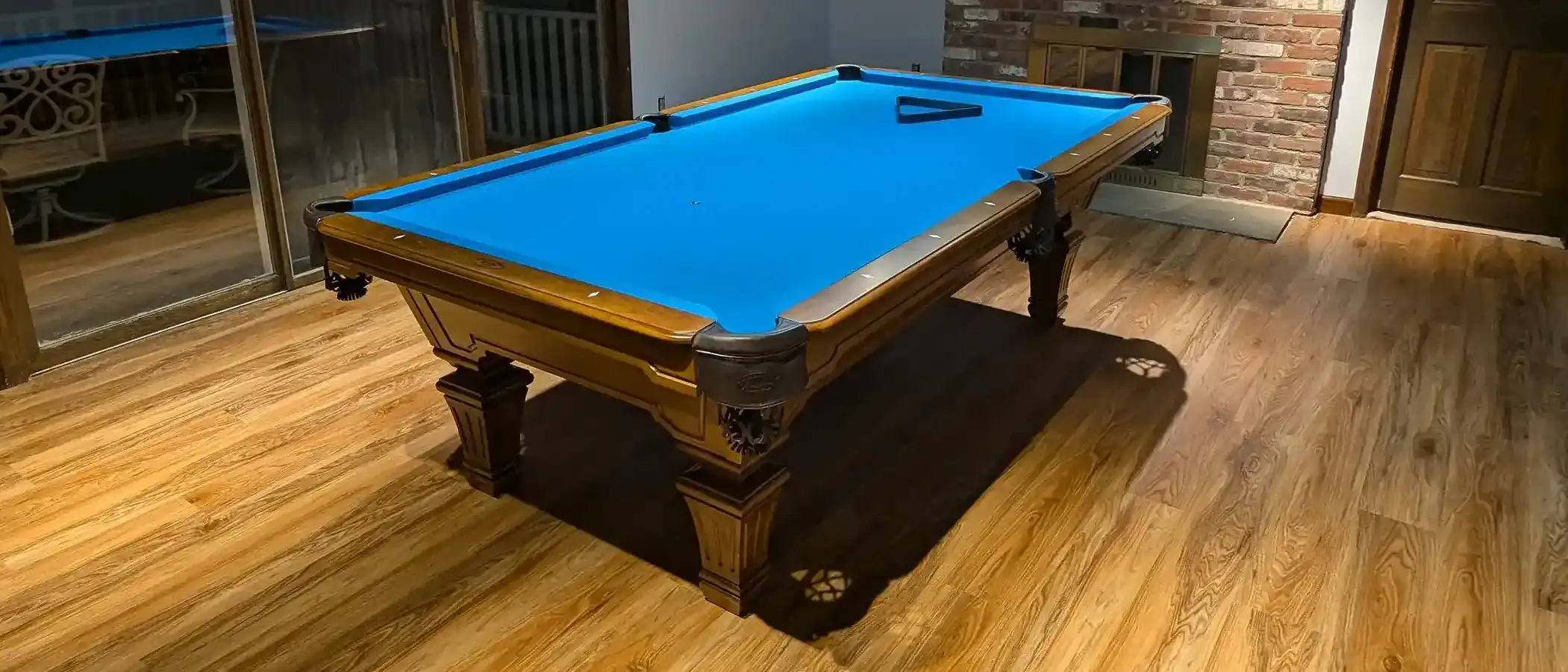 Blue-felt pool table with carved wooden legs on hardwood floor in a cozy game room, near brick fireplace and sliding glass doors, evening lighting.