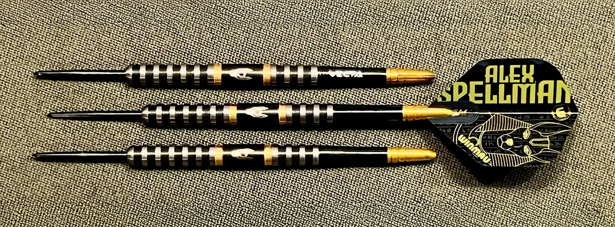 Three black and gold darts with an Alex Spellman branded dart flight. They feature a unique grip pattern and gold tips.