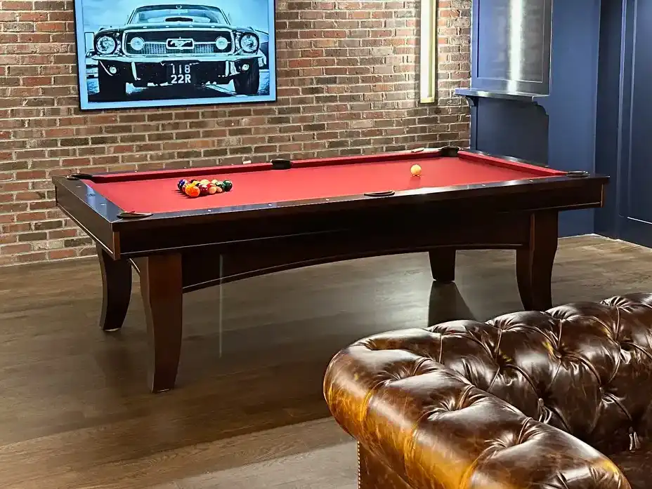 Vintage-style game room with a dark wood pool table featuring red felt, brick accent wall, classic car photo, and tufted leather sofa in front.