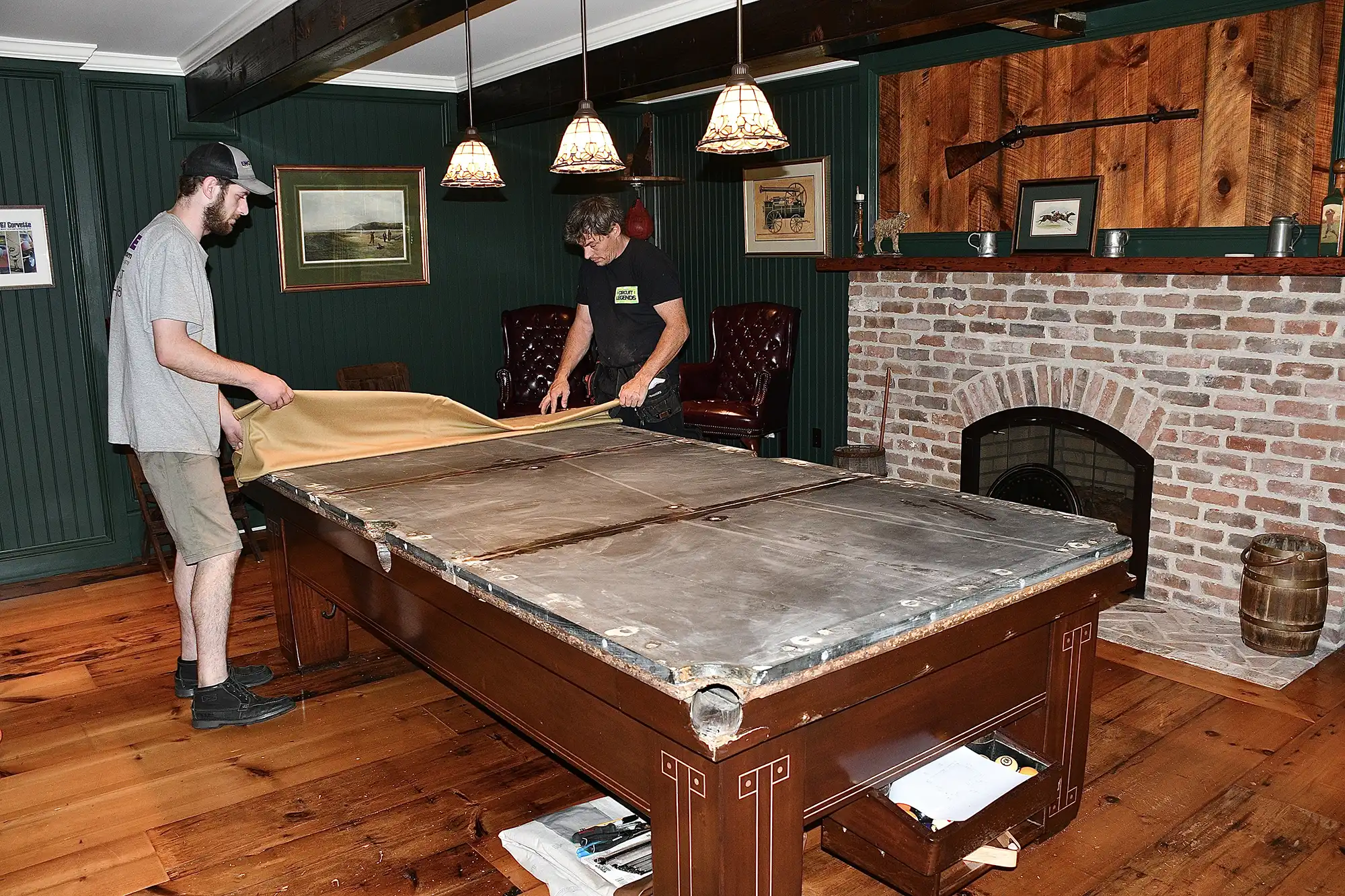 Two people are stretching new felt over a pool table being restored in a cozy game room with wood floors, green paneling, and a brick fireplace.