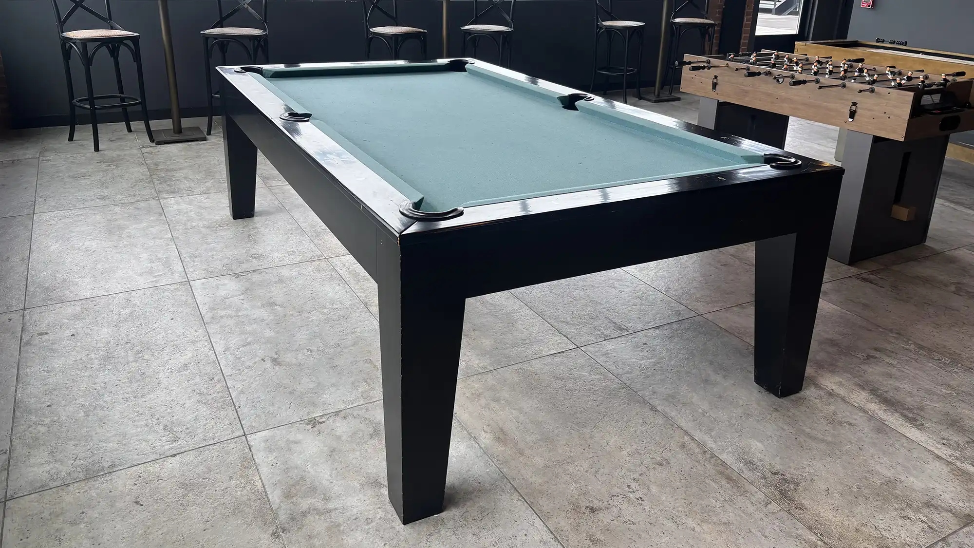Before image of a black wood pool table with light green felt, sleek modern design, and dark flooring in a game room with a foosball table.