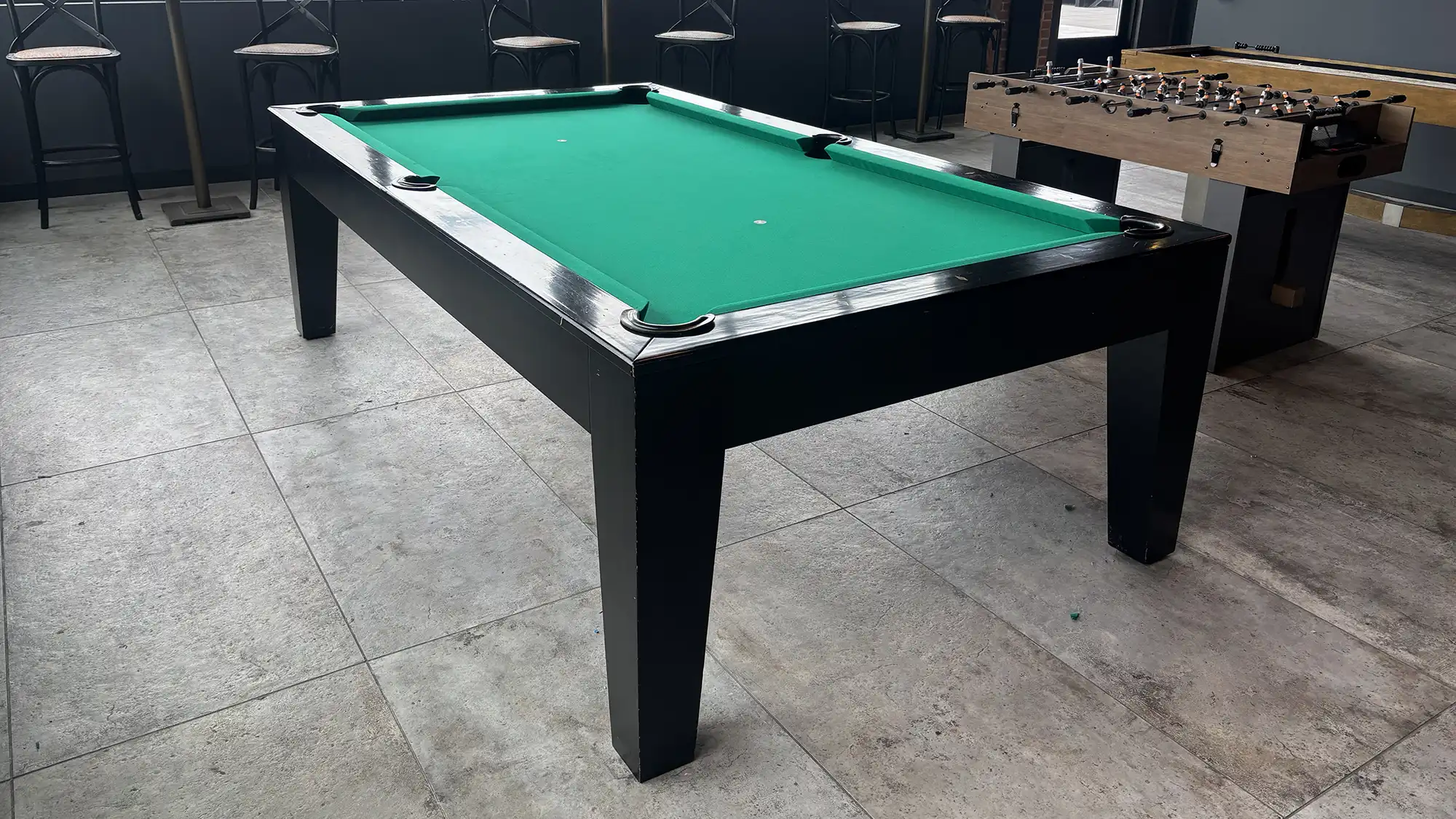 After image of a stylish black wood pool table with vibrant emerald green felt in a modern game room. Features an adjacent foosball table and bar seating.