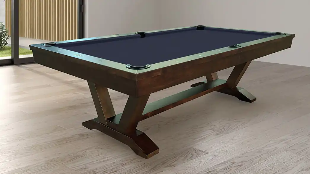 A dark wood pool table with navy blue felt and modern X-shaped legs stands in a bright room with light wood floors and a sliding glass door in the back.